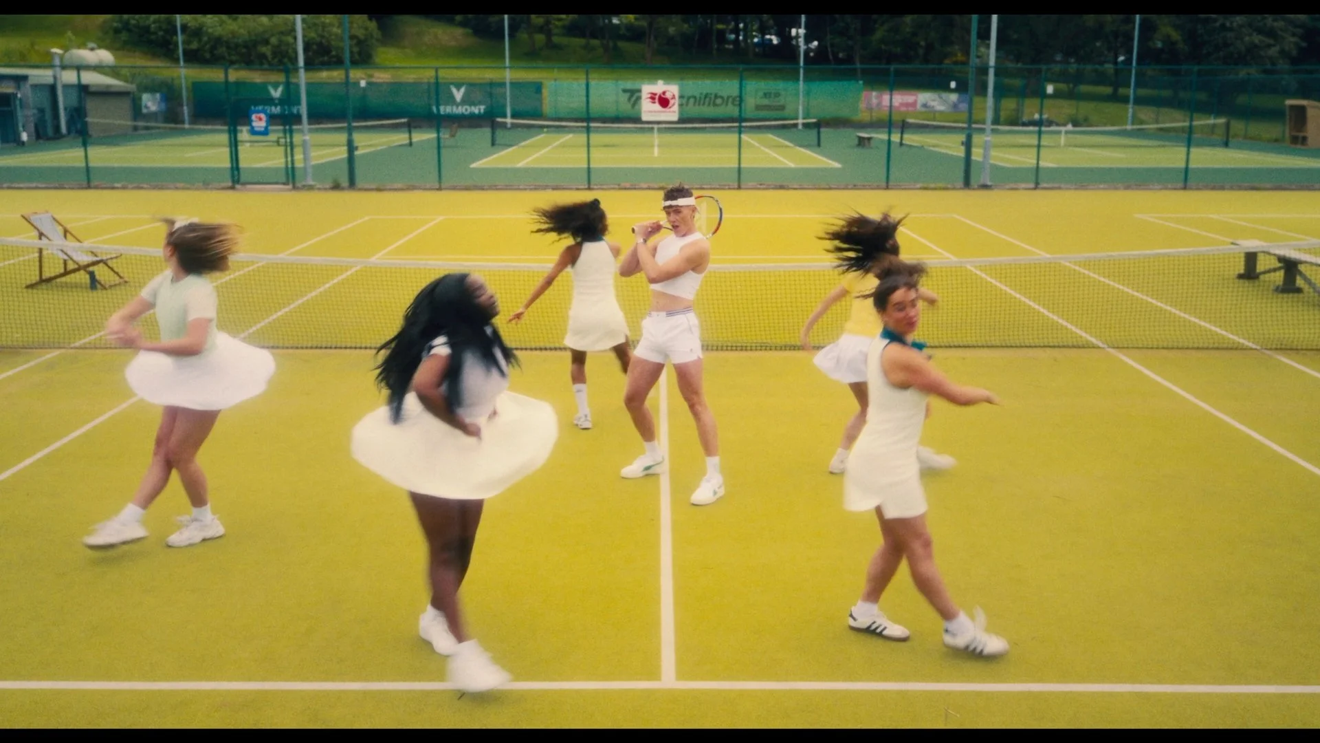 A man dressed in tennis attire, holding a tennis racket and wearing a tennis headband, stands on a tennis court surrounded by five women wearing white dresses, with some spinning or dancing, during daytime.