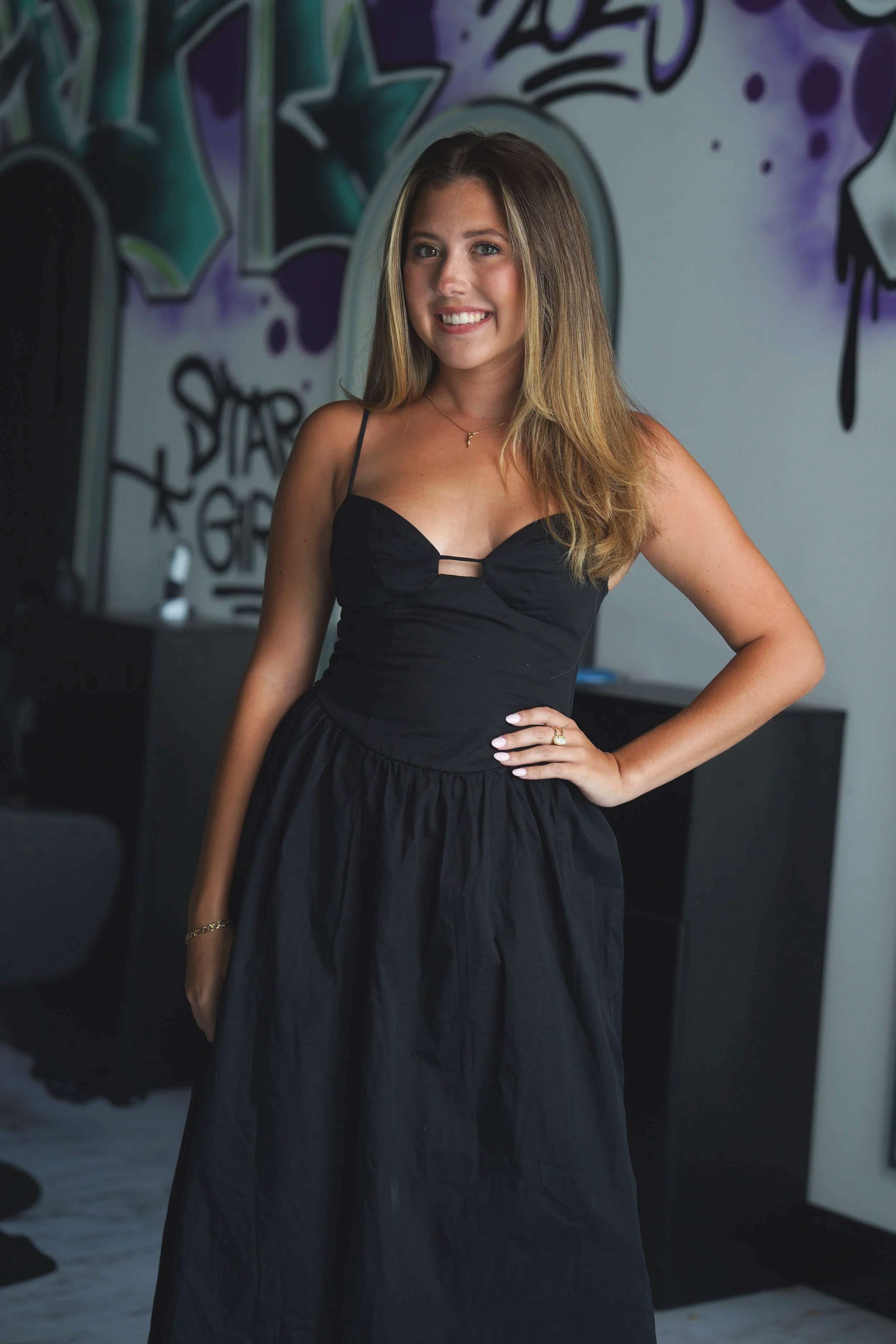 A young woman with long wavy brown hair, wearing a black blazer, black dress, sheer black stockings, and black high heels, is sitting on a white stool against a plain white background. She is smiling and looking at the camera.