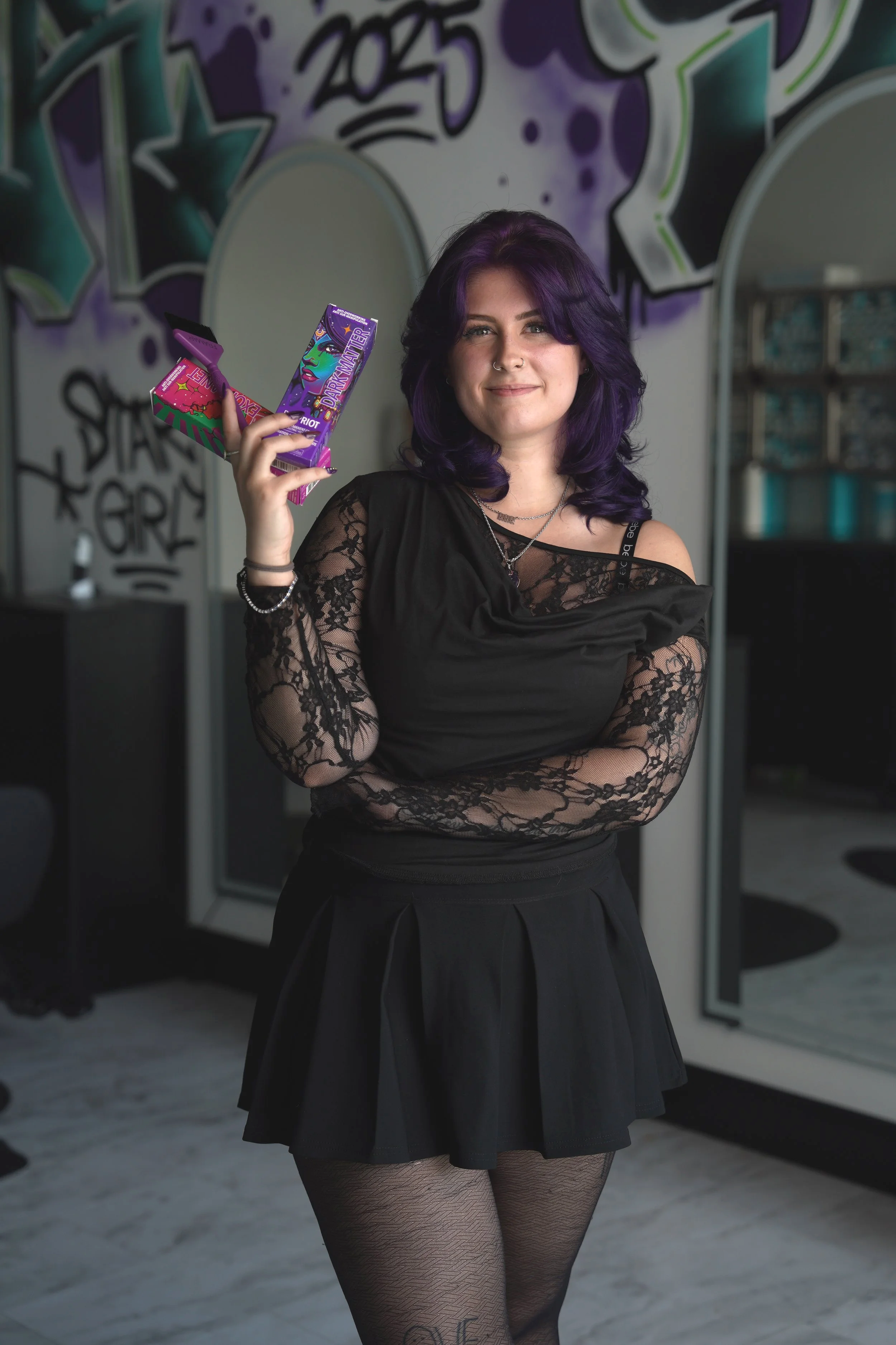 A woman with purple hair and fair skin standing indoors, wearing a black lace long sleeve top, black skirt, and patterned black tights. She is holding a purple box labeled 'Dark Matter' and smiling slightly. The background features a wall with graffiti art and a mirror.