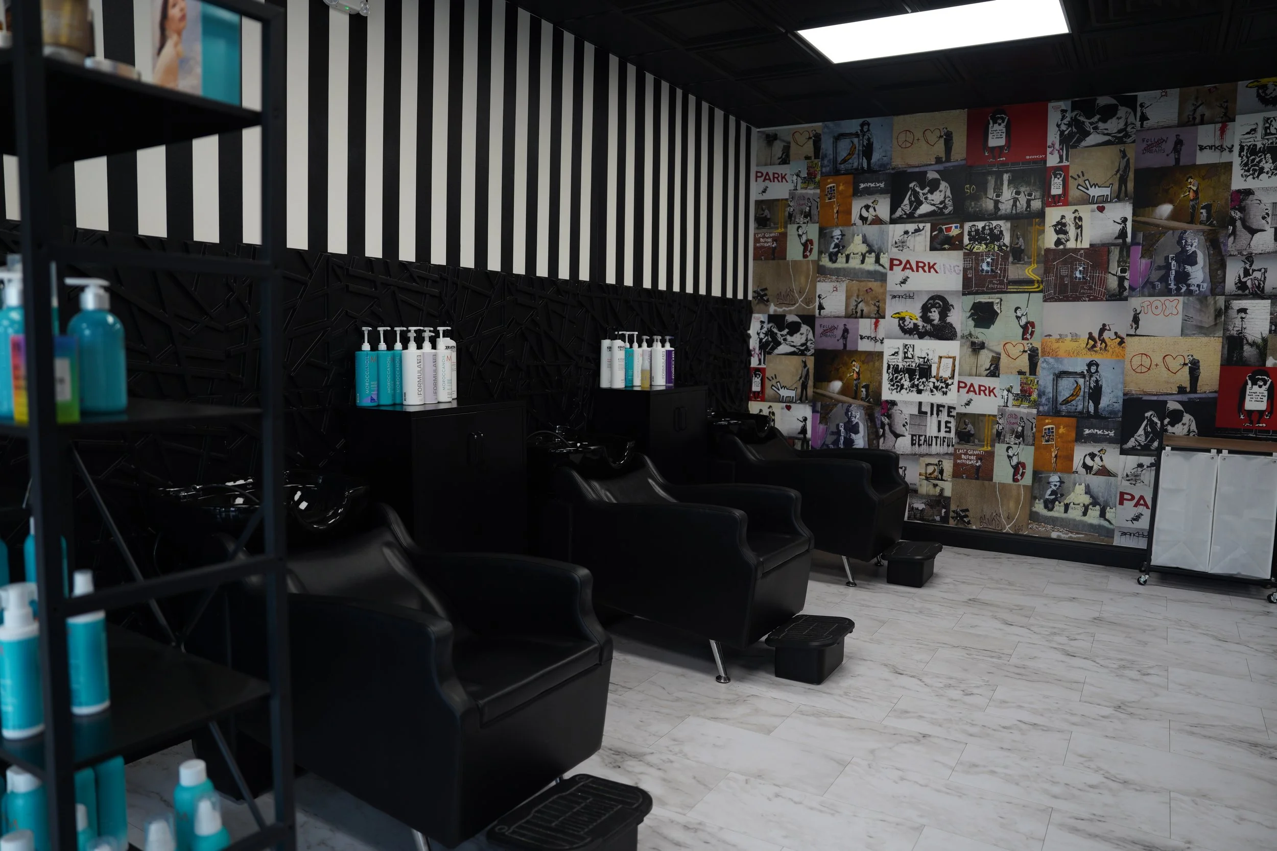 A hair salon interior with black salon chairs, white marble flooring, and a wall covered with colorful street art-style murals, including images of people, peace signs, hearts, and words like 'PARK' and 'LIFE IS BEAUTIFUL.' There are shelves with hai