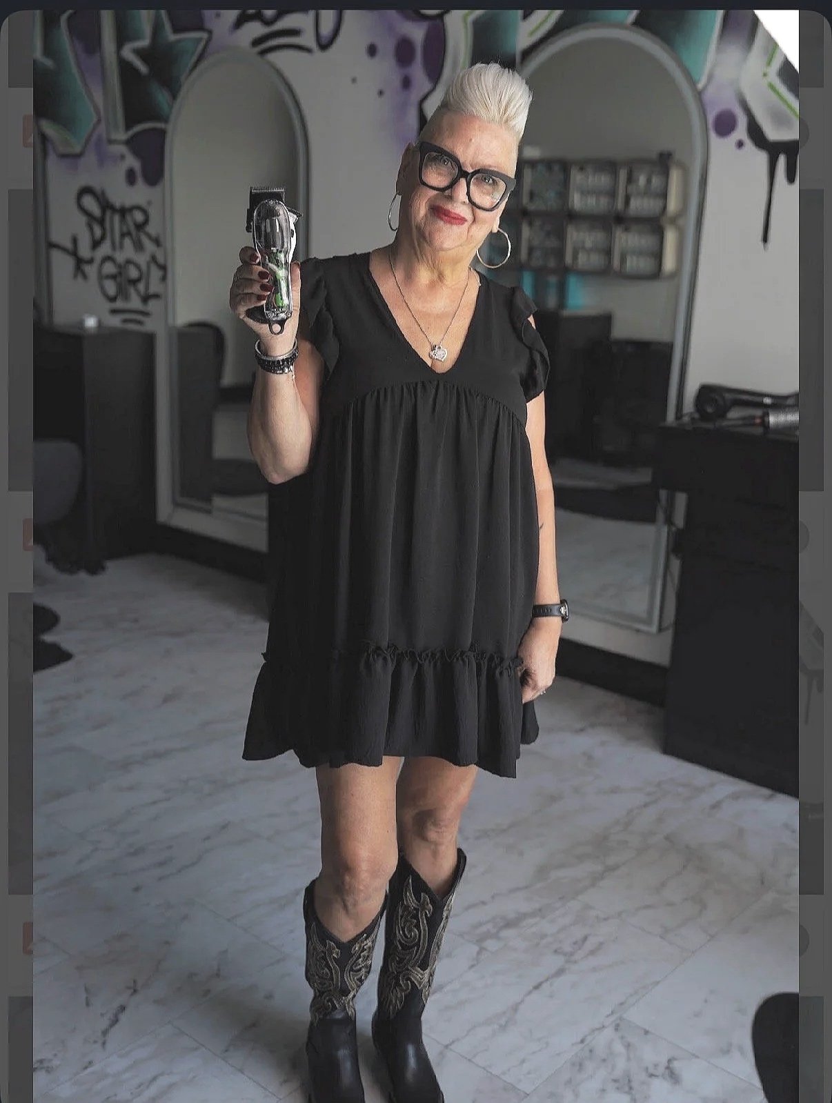An older woman with platinum blonde hair styled in a pompadour, wearing large black glasses, red lipstick, a black off-shoulder dress with a rose pattern, and a silver necklace, sitting against a plain white wall.