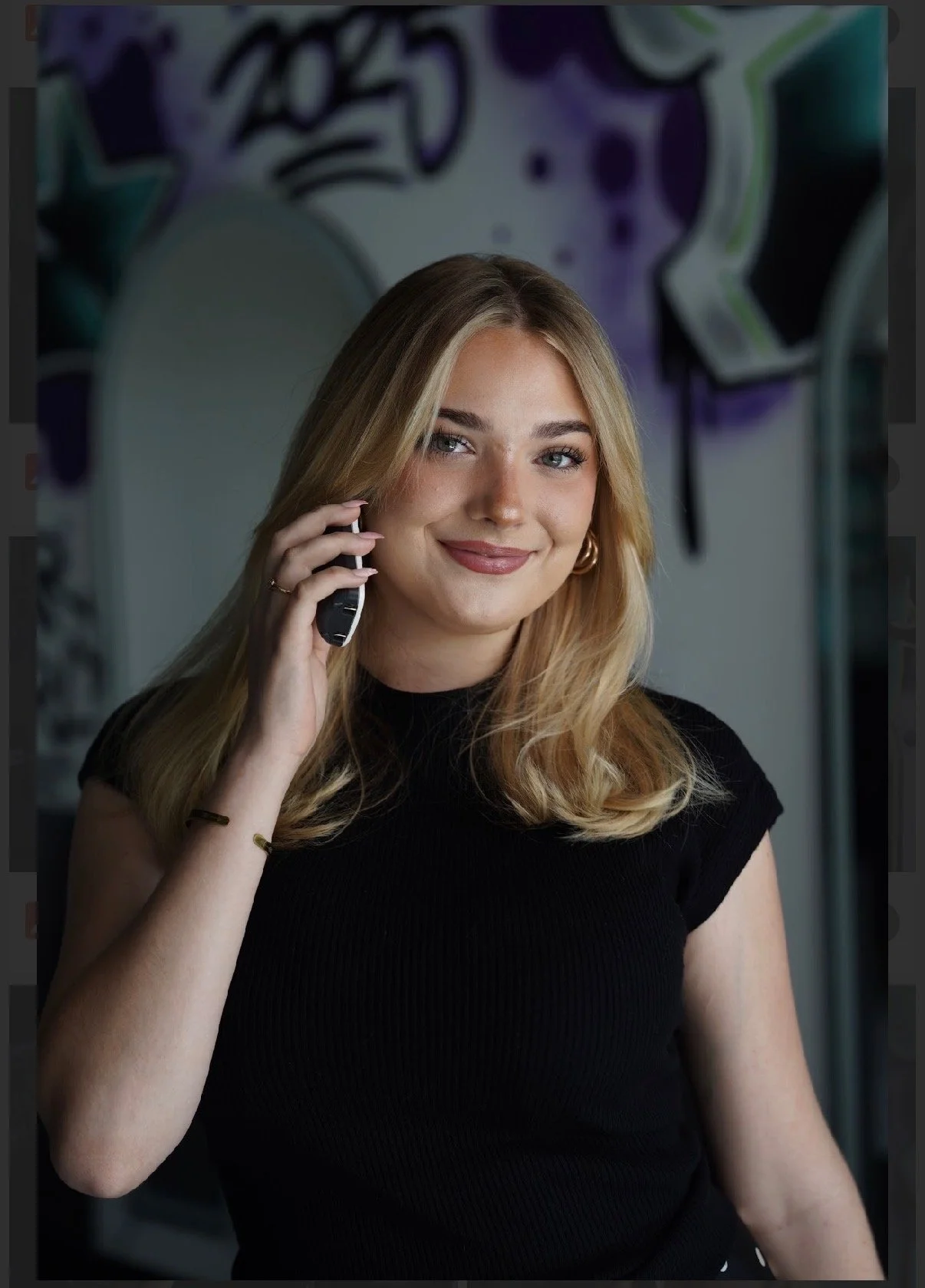 A young woman with shoulder-length blonde hair, wearing a black blazer over a white shirt, posing against a plain white wall.