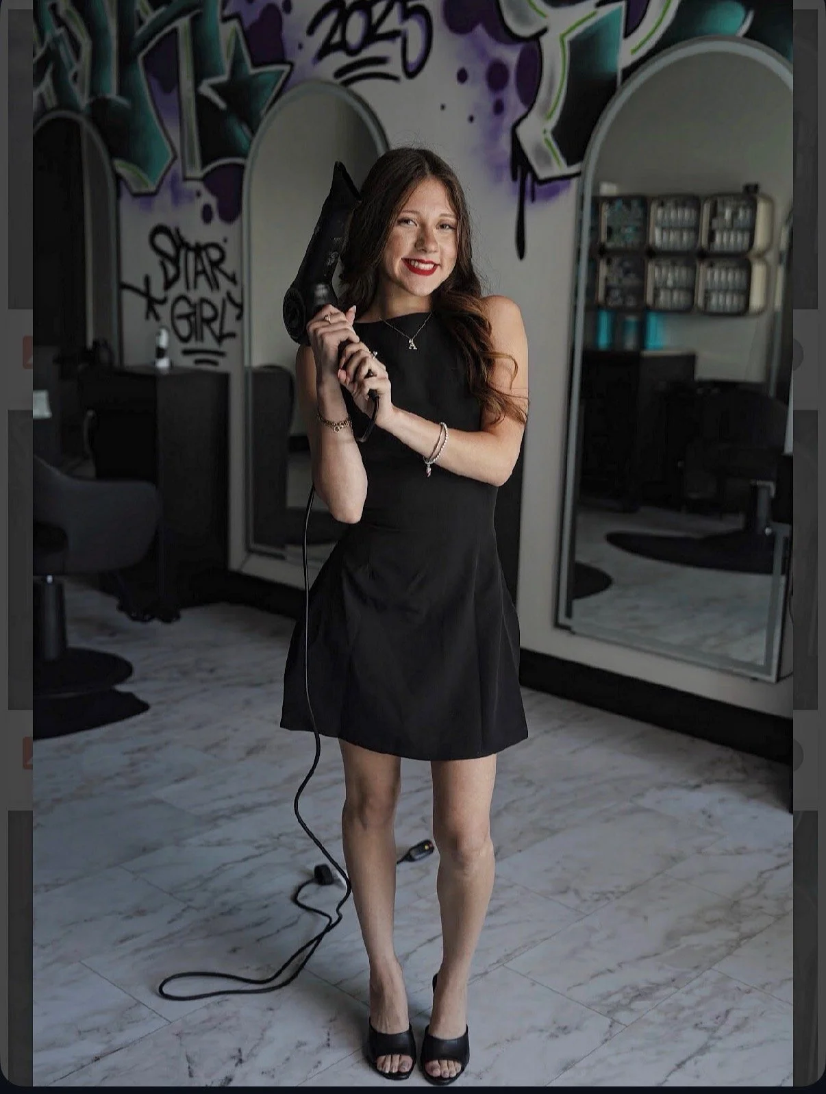 A woman sitting on a stool against a plain white wall, wearing a black long-sleeve top, black pants, black high heels, and accessorized with a necklace, earrings, and a watch.