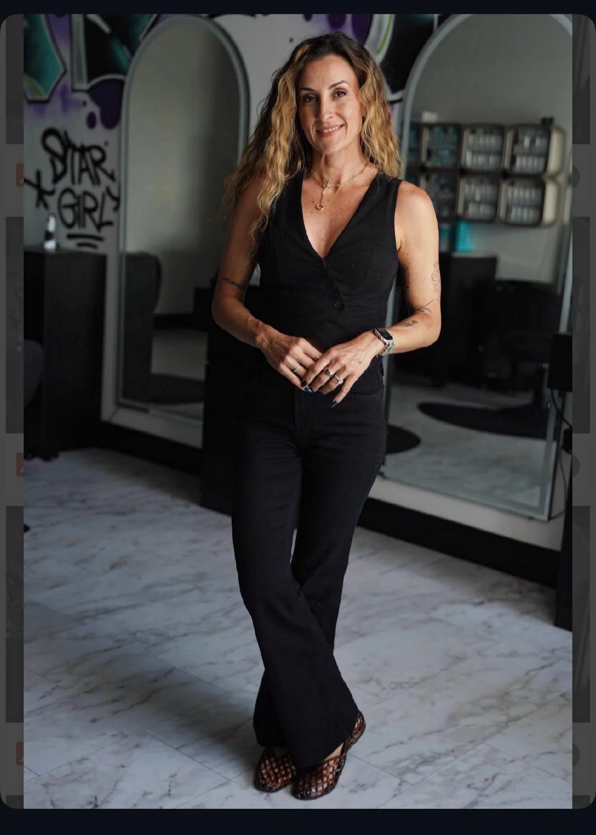 A woman with wavy hair and a leopard headband standing inside a beauty salon, holding two signs that read "ALT AIR SALON" and "BOOK NOW." The background features a collage of black and white photos on the wall and shelves stocked with hair care products.