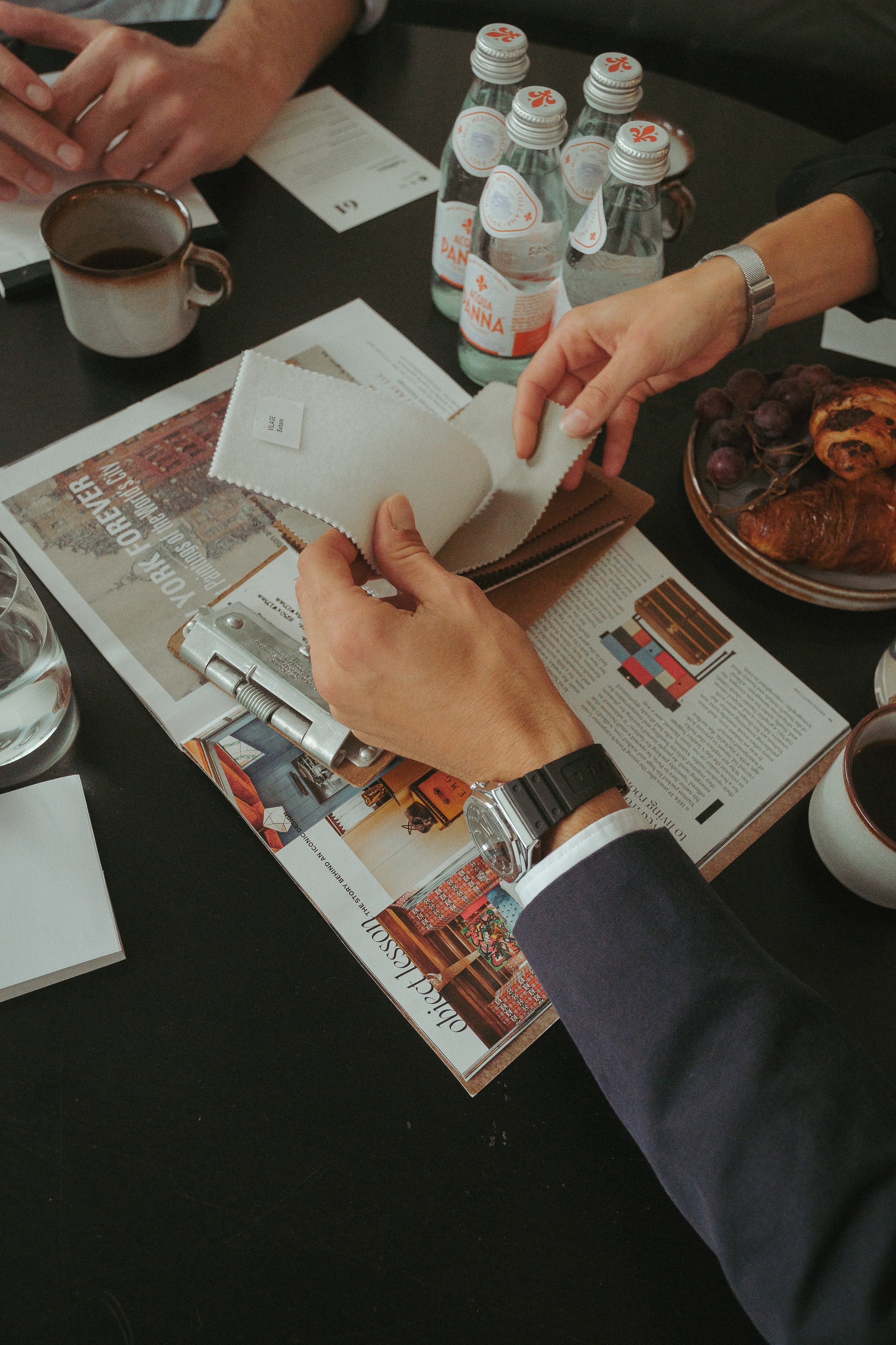 Meeting table with water bottles, coffee, and magazines; two people are handling fabric samples.