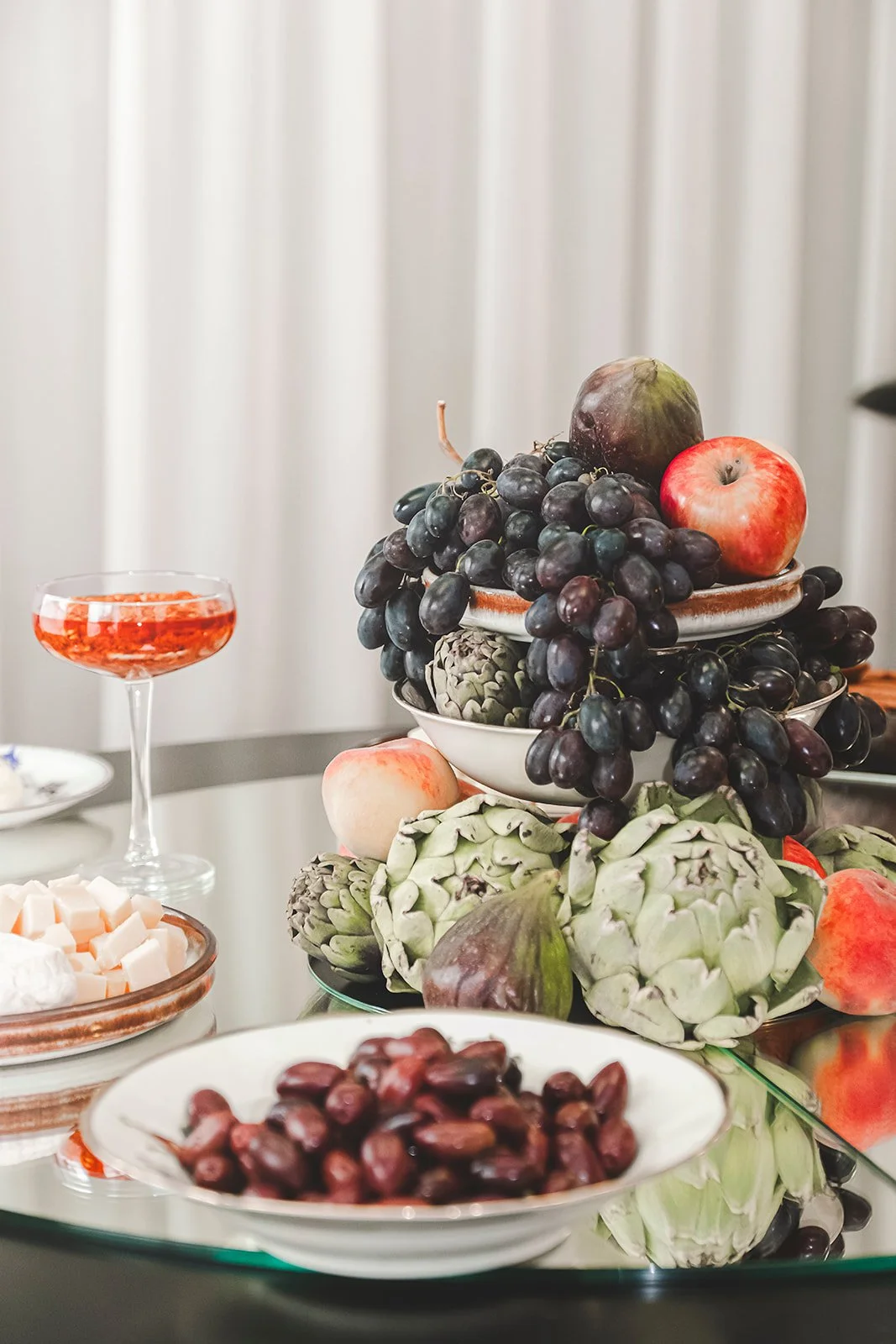 A table with various fruits including grapes, apples, peaches, figs, artichokes, and a bowl of red beans, with a glass of red drink.