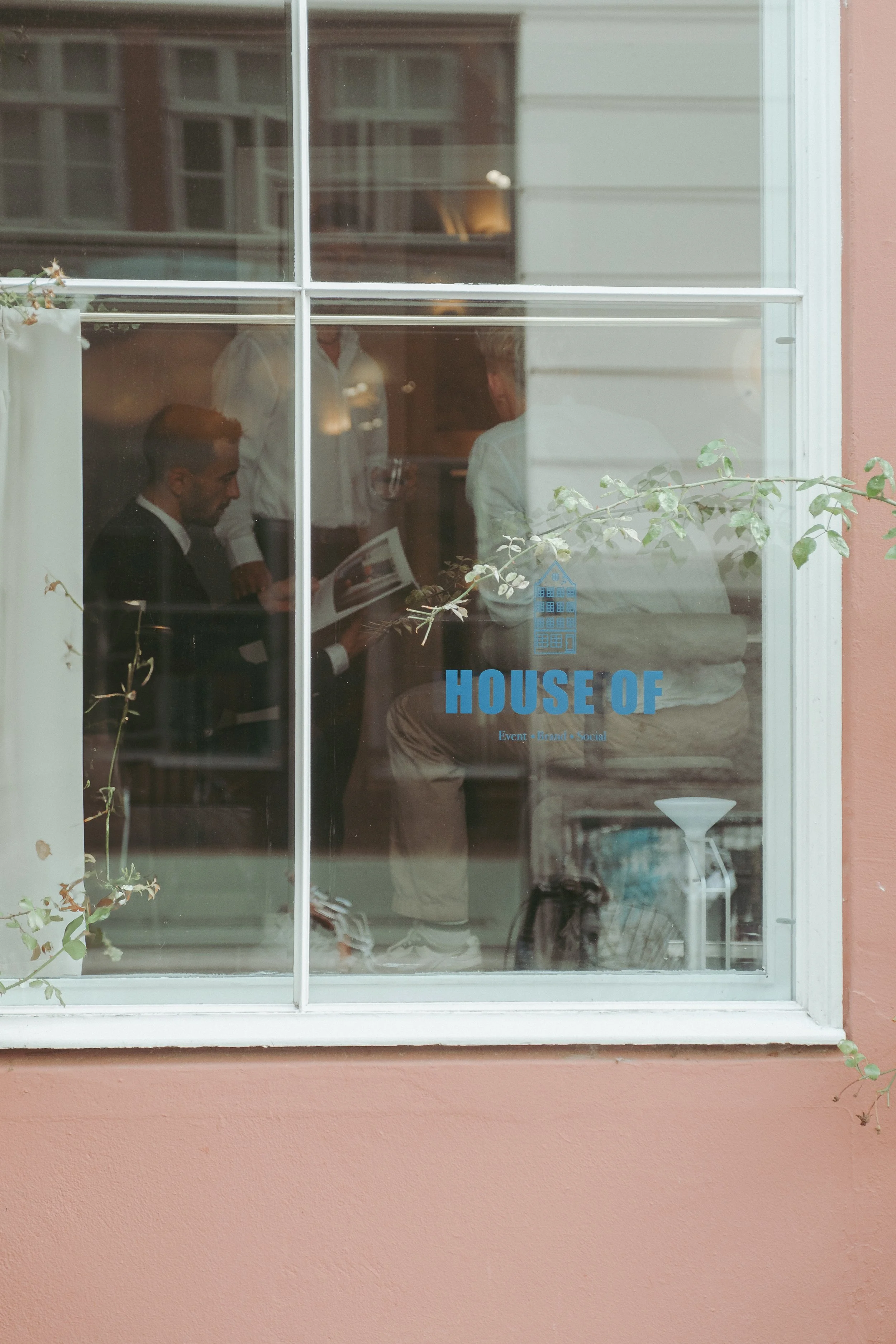View through a window with a blue logo that says 'HOUSE OF' and people inside appearing to be having a meeting or discussion.