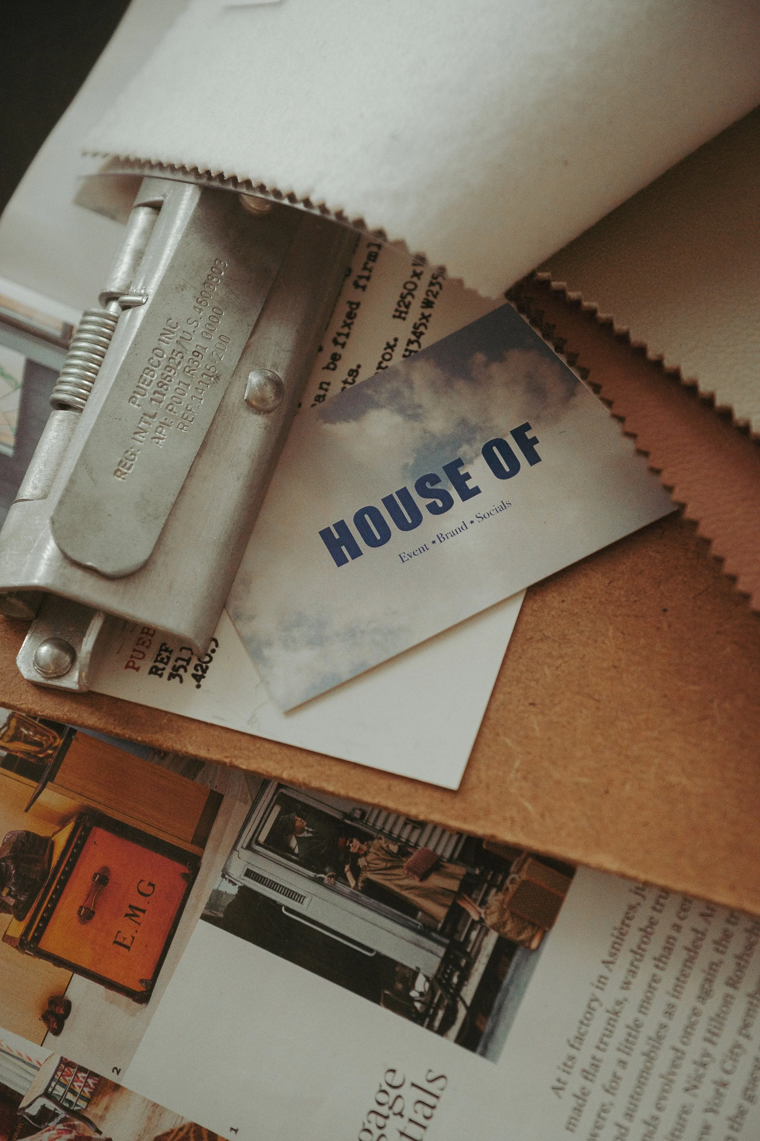 A close-up view showing a metal stapler, a business card that reads 'HOUSE OF', and some printed materials on a wooden surface.