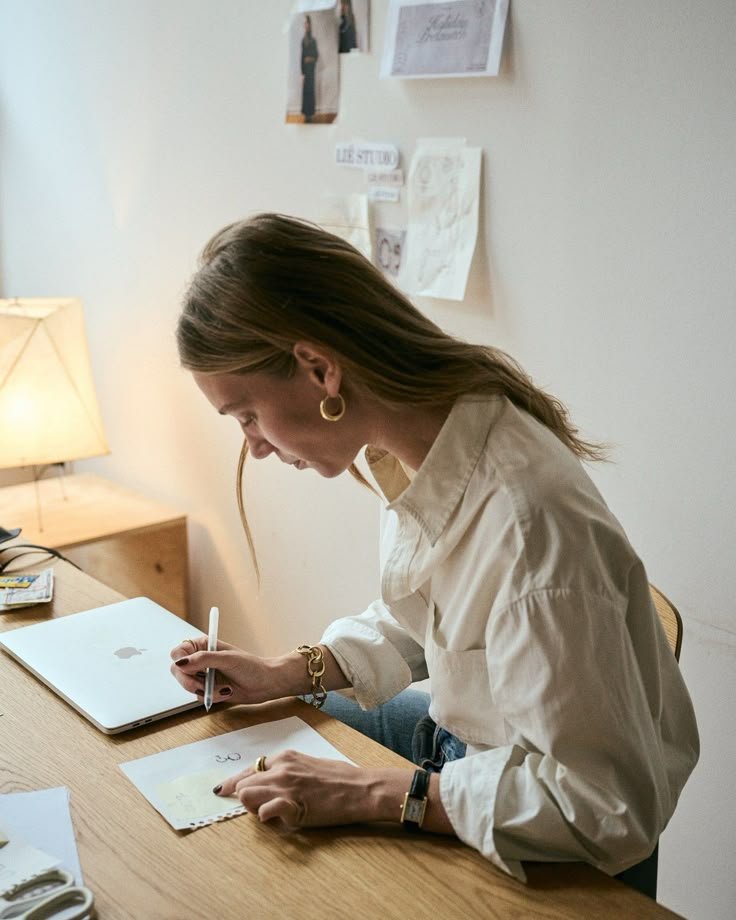 A woman with long brown hair and hoop earrings is sitting at a wooden desk, writing on a piece of paper with a white pen. She is wearing a beige button-up shirt and has a watch on her left wrist. Next to her is a closed silver MacBook. The background includes a beige wall with various notes and pictures pinned up, and a lamp providing warm lighting.