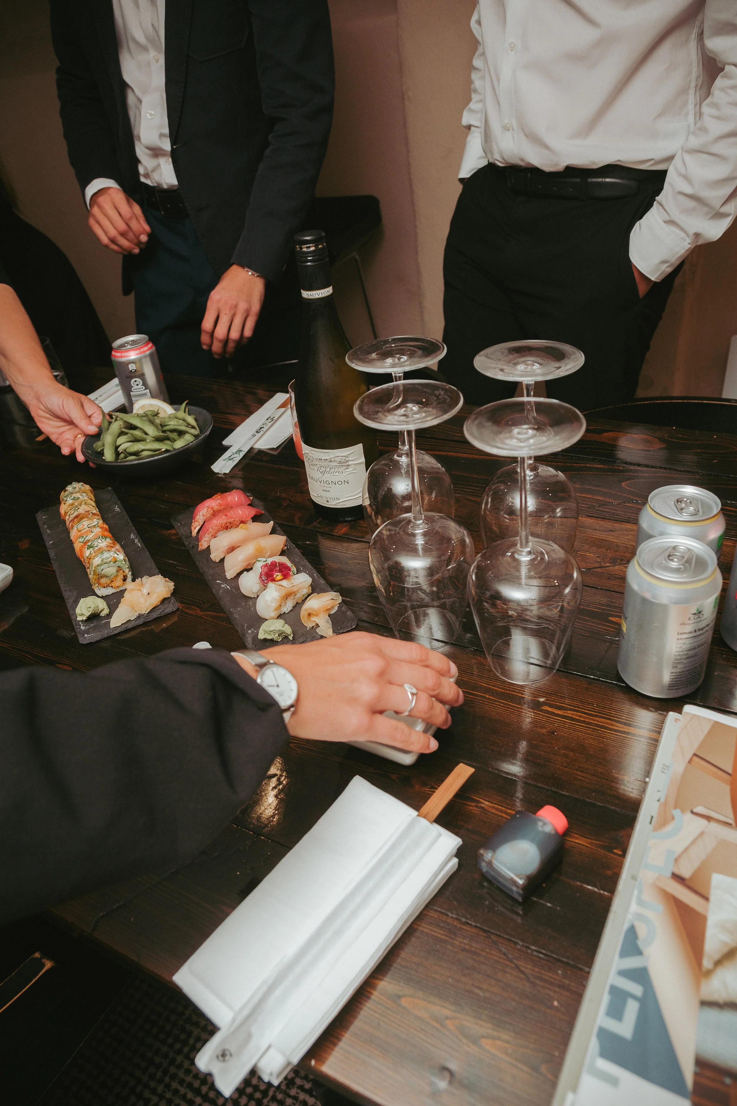 A table set with sushi, sake, canned drinks, wine glasses, and edamame, with several people standing around.