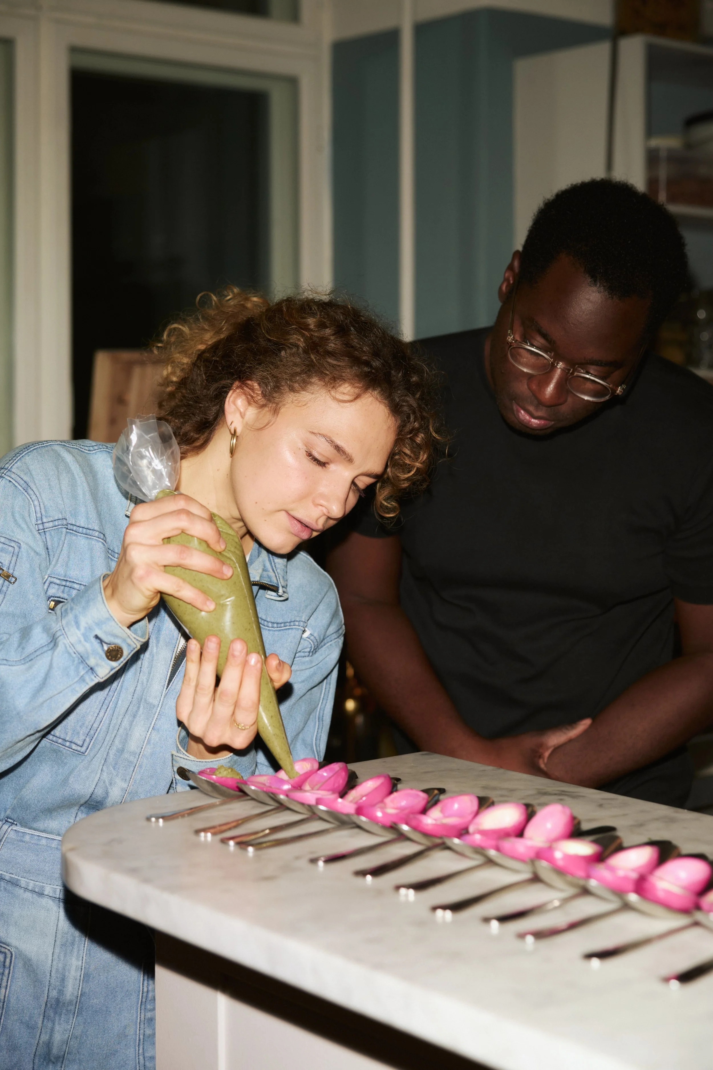 A woman and a man decorating pink macarons on a marble countertop in a kitchen.