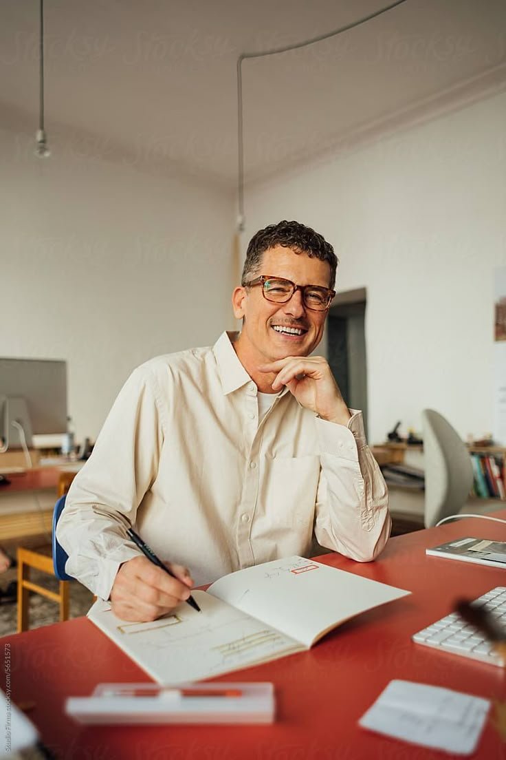 A man with glasses sitting at a desk, smiling, holding a pen, with papers, a keyboard, and a computer monitor in his workspace.