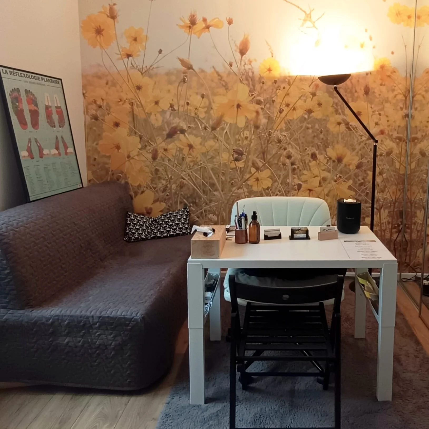 Une pièce avec un mur à motif floral jaune, un canapé gris avec un coussin noir et blanc à motifs, une table blanche avec des fournitures de bureau, une chaise noire, une lampe de sol et un poster de réflexologie plantaire.