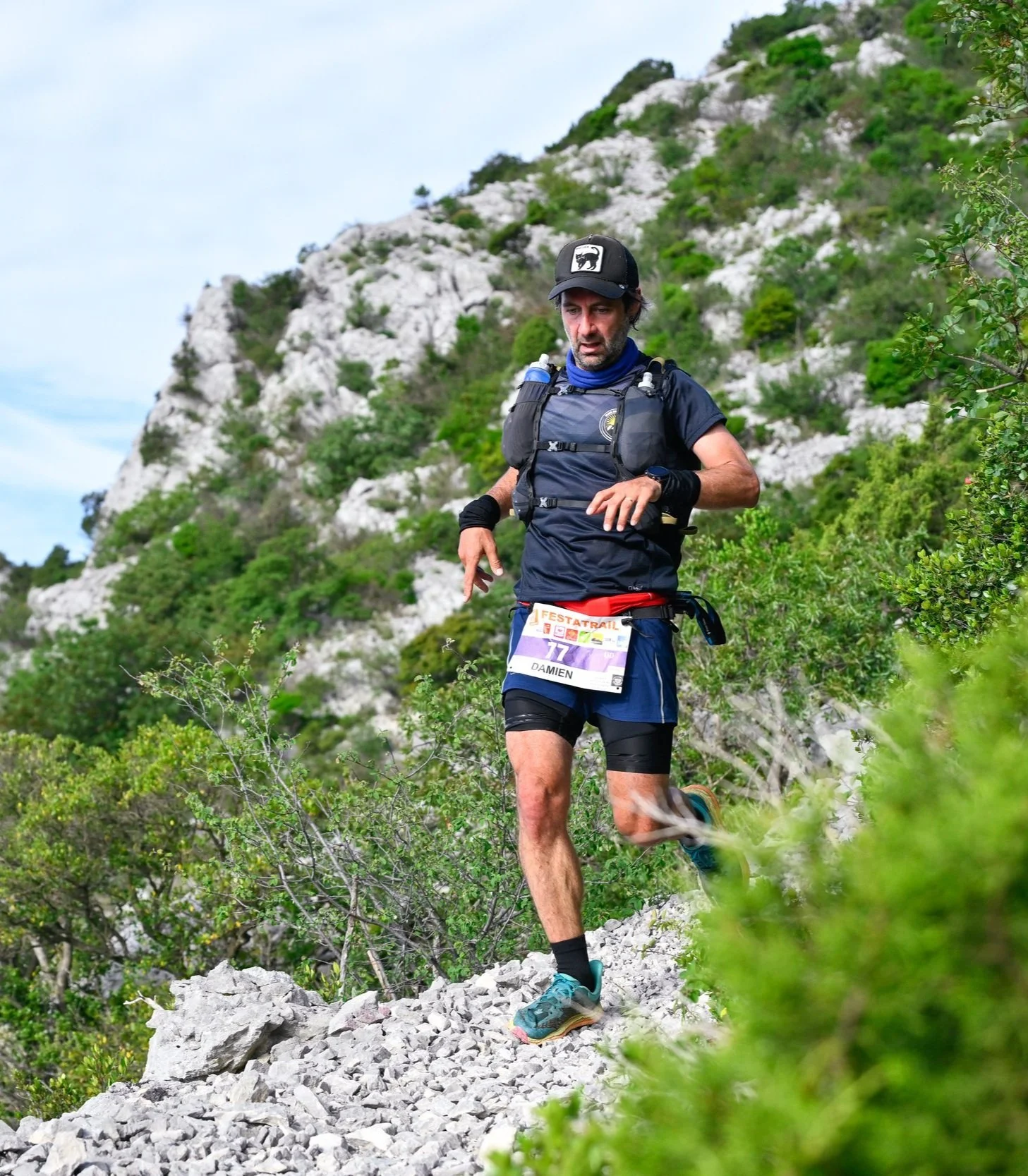 Un homme courant en trail dans un environnement montagneux avec des rochers et des végétations verdoyantes, portant un sac à dos, un maillot noir, un chapeau, et un numéro de course affiché avec le nom Damien.