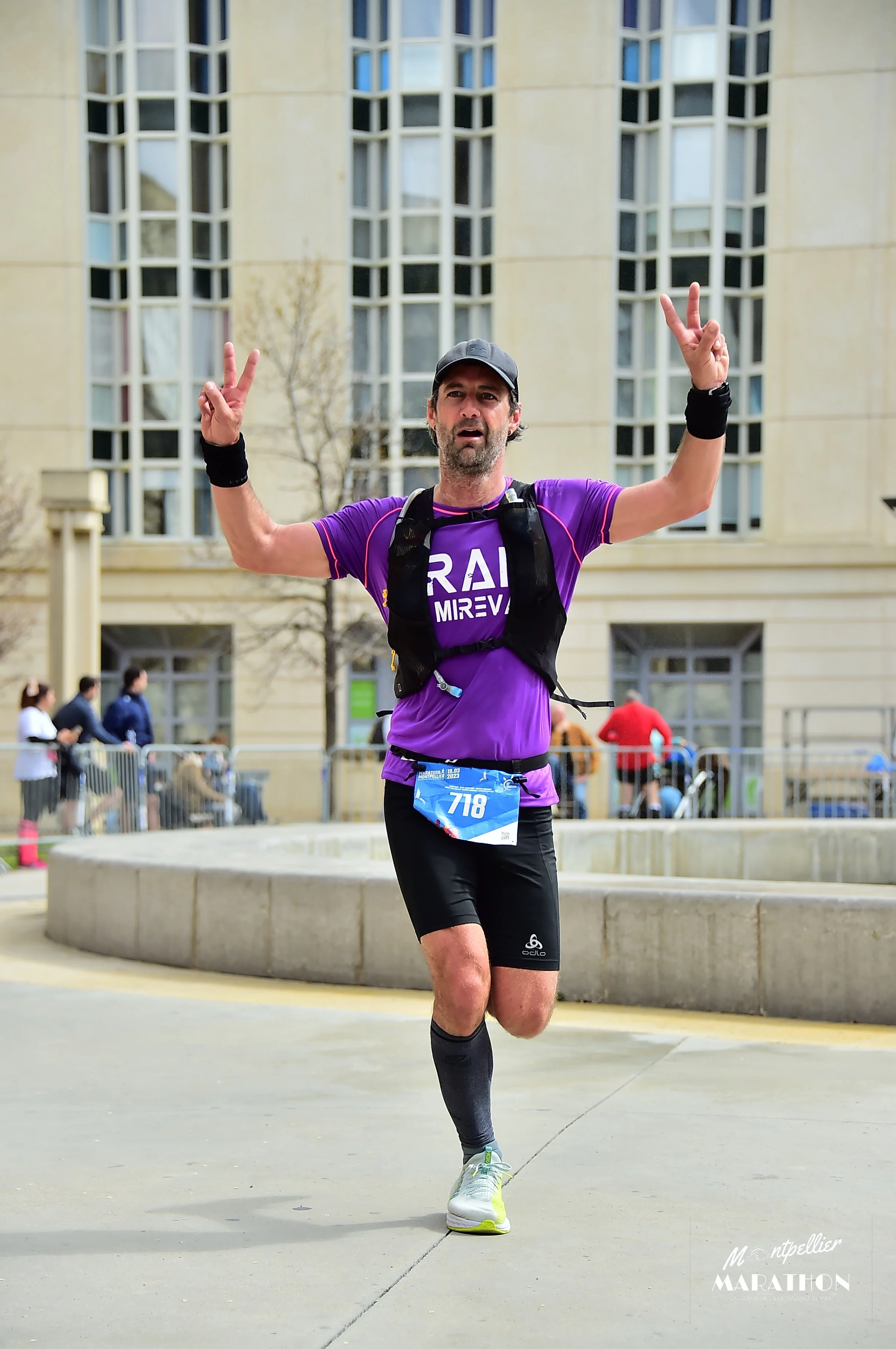 Un homme court lors d'un marathon, portant un t-shirt violet, un short noir, une casquette, et fait un signe de paix avec les deux mains. Il y a un bâtiment en arrière-plan et d'autres personnes à l'arrière.