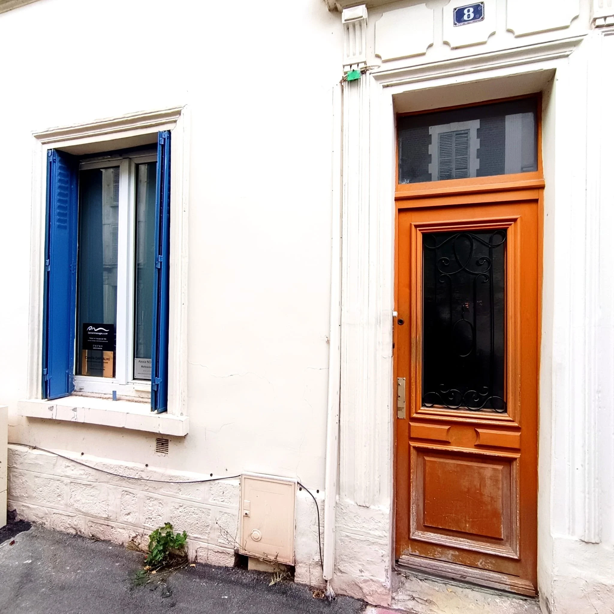 Façade d'un bâtiment avec deux fenêtres et une porte, la fenêtre de gauche a des volets bleus et la porte est en bois avec un cadre orné, numéro 8 en haut à droite.