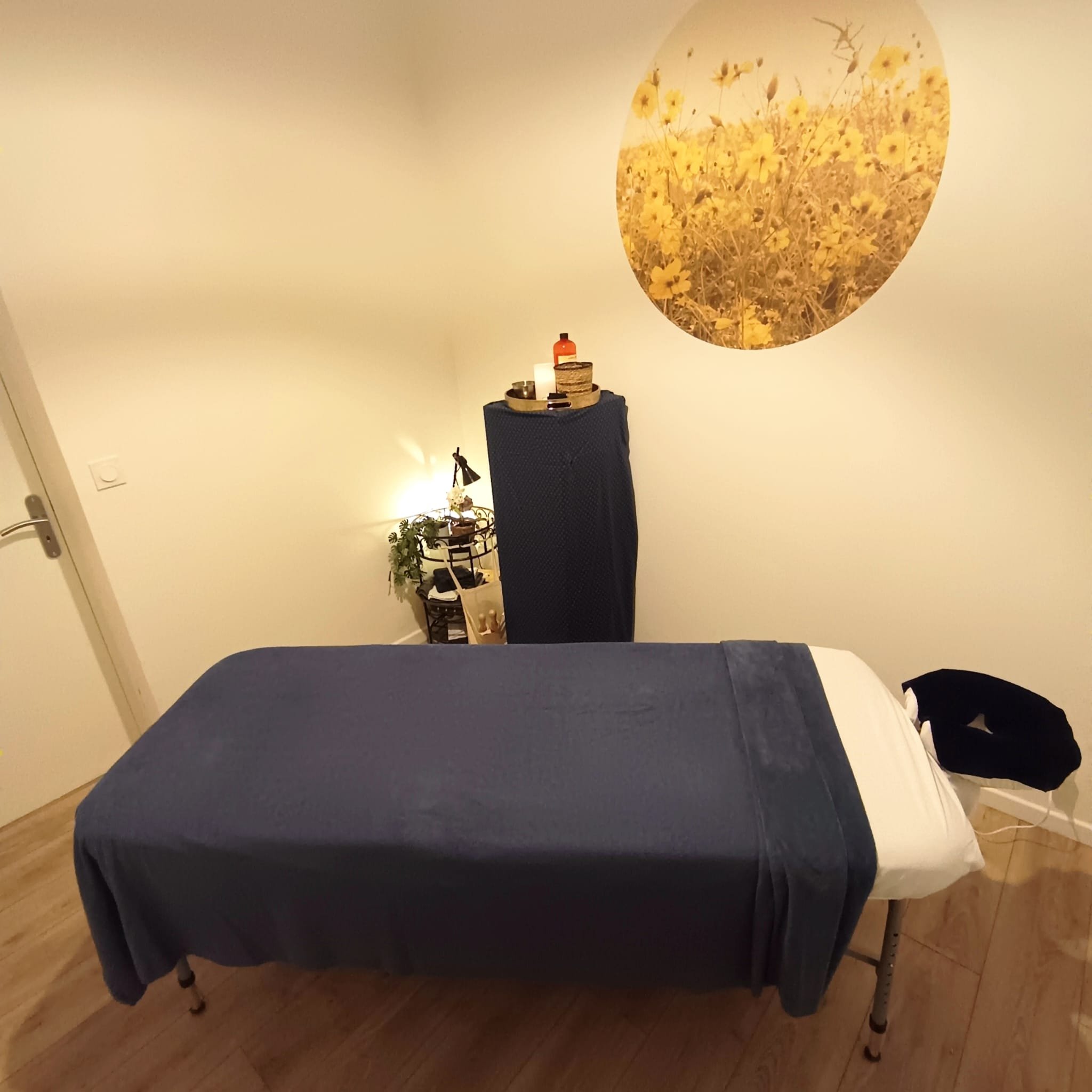 Table recouverte d'un tissu noir dans une pièce avec une décoration florale dans un pot en céramique, un livre et une bouteille orange, avec un mural en ronde avec une image de champs de fleurs jaunes en arrière-plan.
