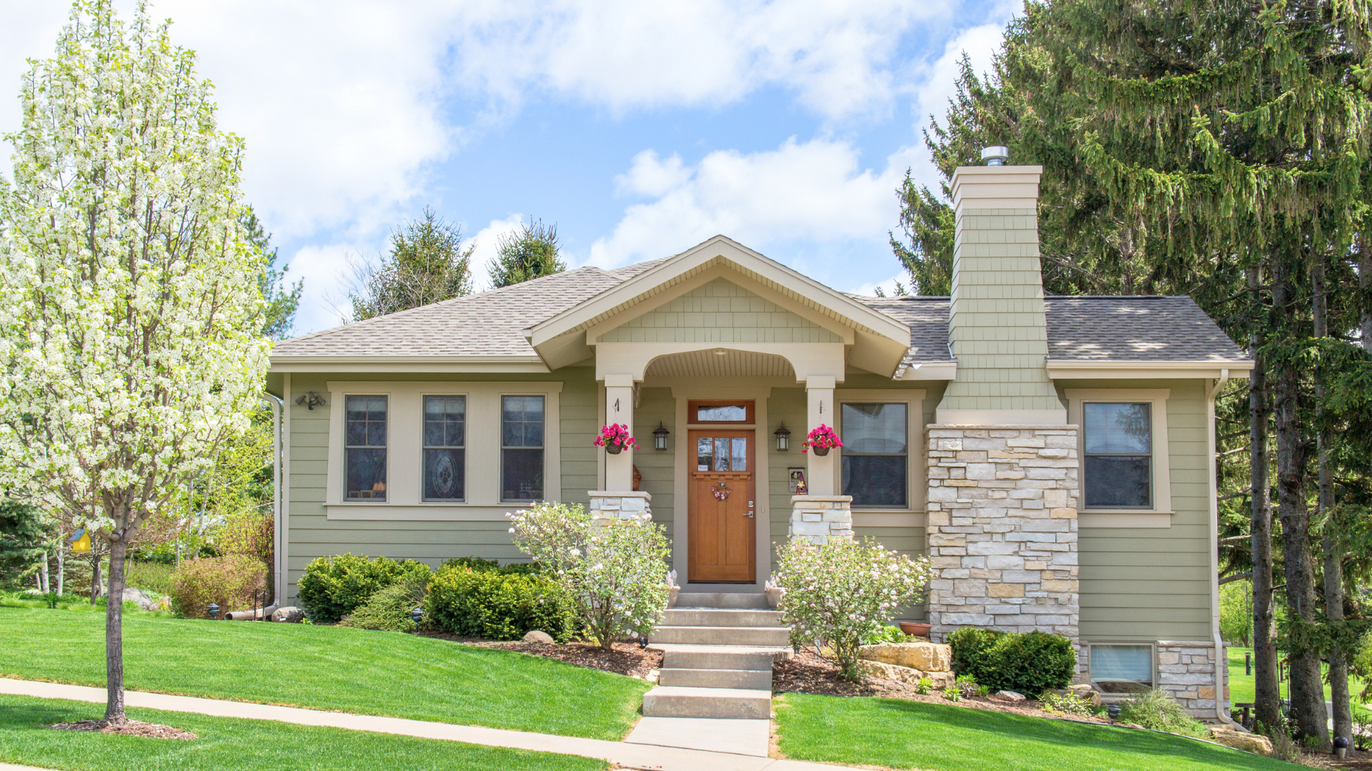 Single-story green house with a gabled roof, stone chimney, and wooden front door. The yard is landscaped with shrubs, a blooming tree, and a manicured lawn. Decorative red flowers hang near the entrance.