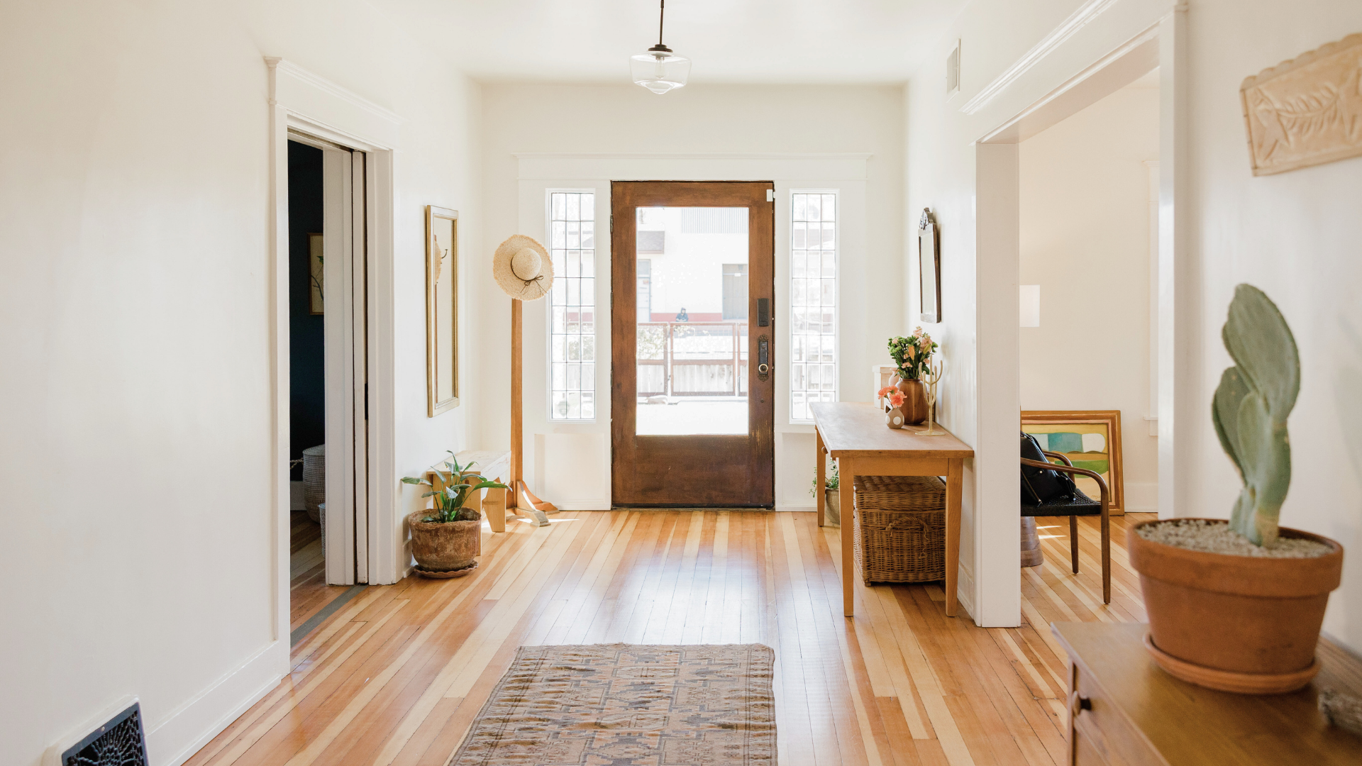 Bright, minimalist hallway with light hardwood floors, a wooden bench, potted plants, a hat stand, and a front door with side windows.