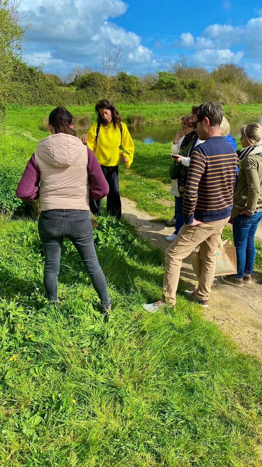 Groupe de personnes discutant dans un champ herbeux, près d'un étang, sous un ciel bleu partiellement nuageux.