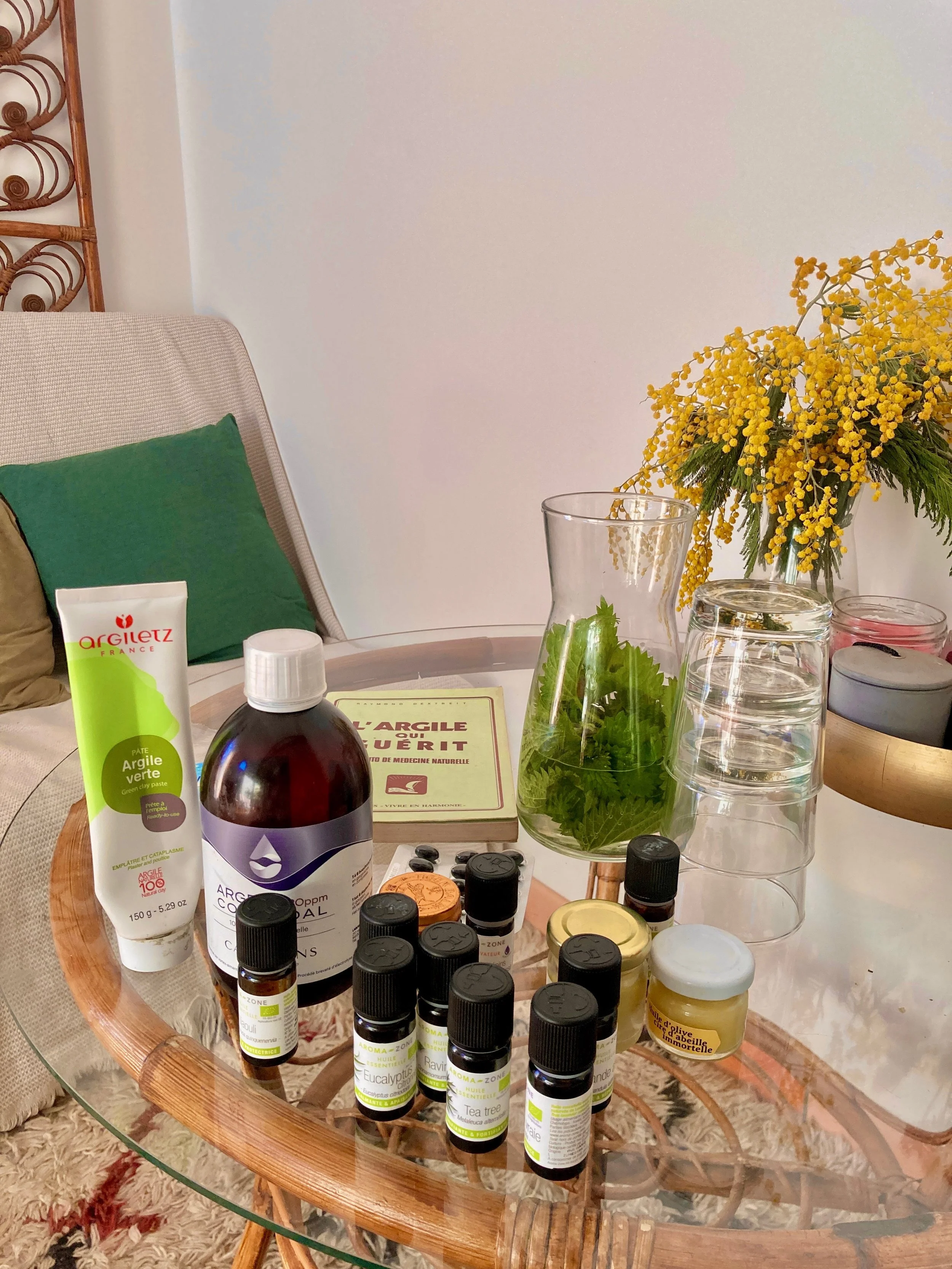 Table en verre avec huiles essentielles, argile verte, bouteille marron, livre, vase avec feuilles vertes, deux verres empilés, et fleurs jaunes.