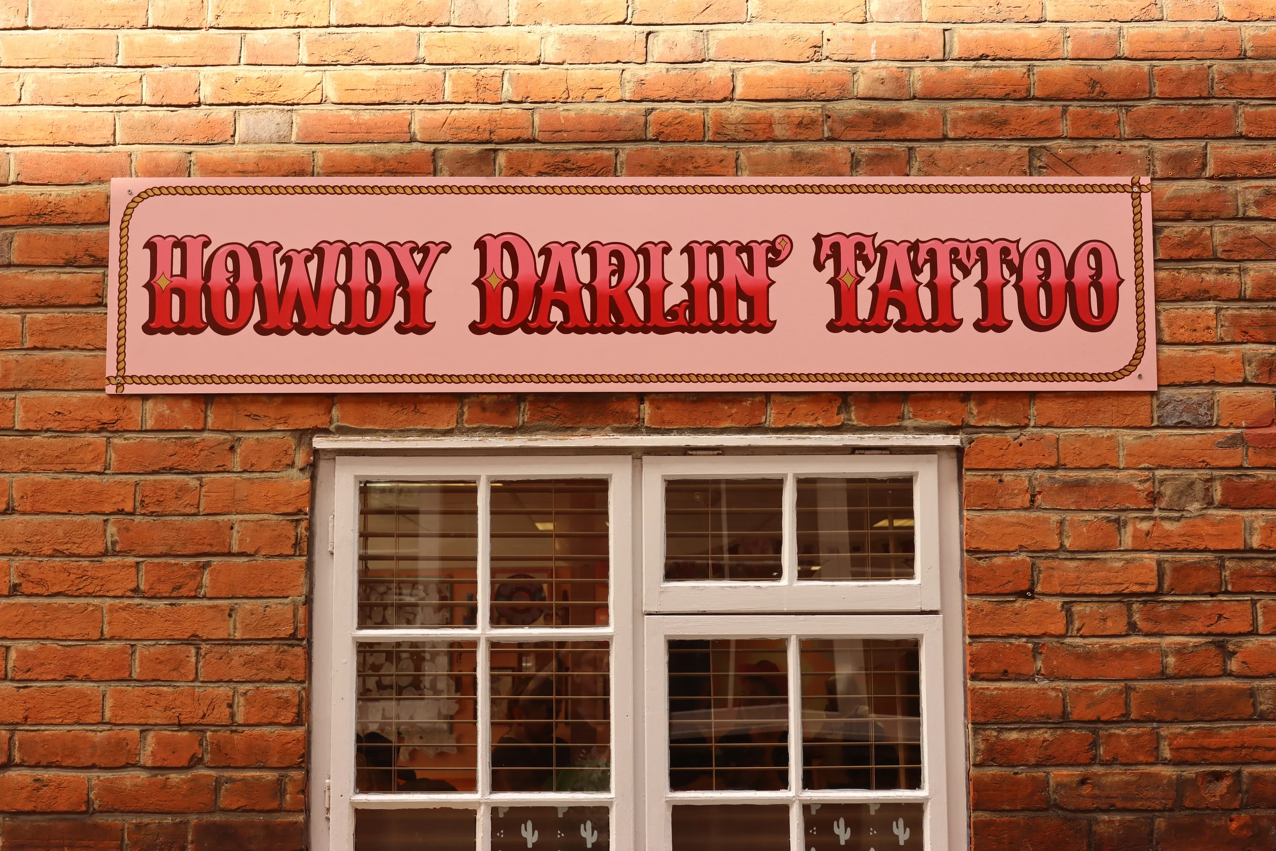 A photograph of Howdy Darlin's rectangular exterior sign on a brick wall. The background is light pink, and the lettering is a medium pink to maroon gradiant. There is a boarder around the words is a light brown rope design.