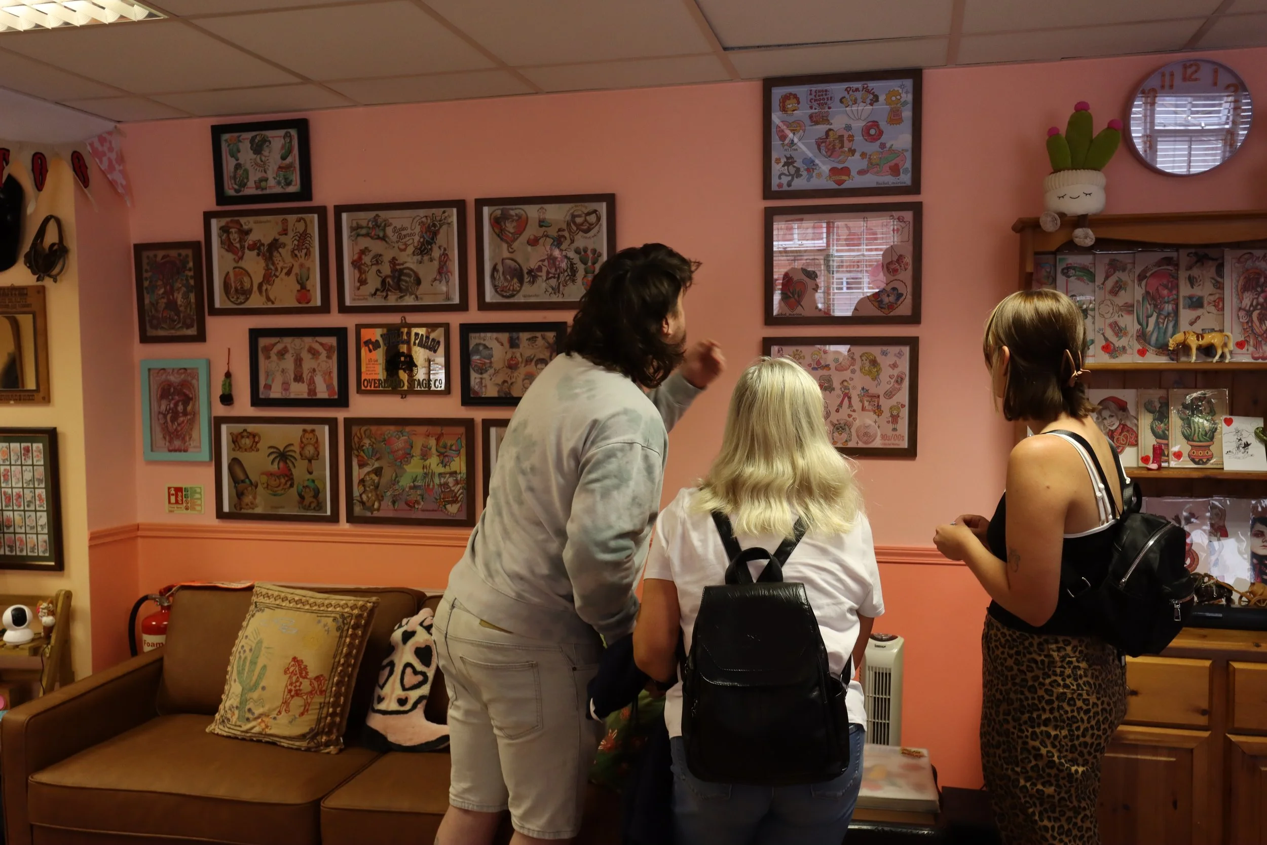 Three people are in the Howdy Darlin' waiting area. They face away from the camera, looking at a wall that has many framed flash sheets on it. The tallest one is in a grey green tie-dye shirt, and is pointing out an image. There is also a brown couch