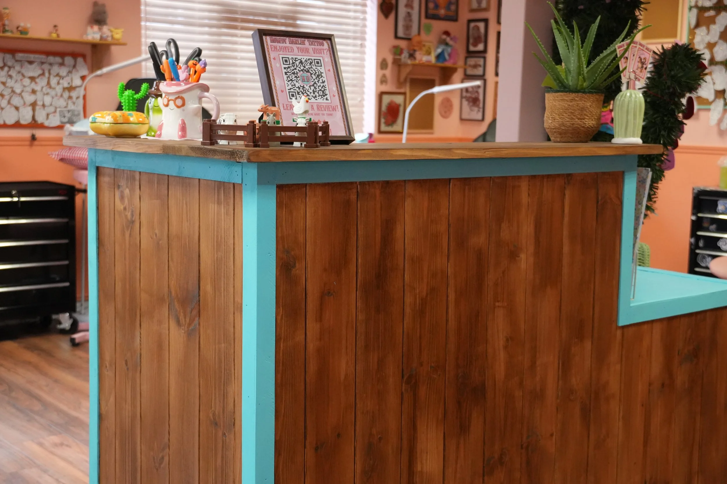 A photo of Howdy Darlin's front desk. It is wooden with a medium-dark finish. The edges are painted a bright turqouise. On the desk is a framed QR code for reviews, a set of Duplo farm animals, a ghost mug that has a cowboy hat on, and a frog cowboy