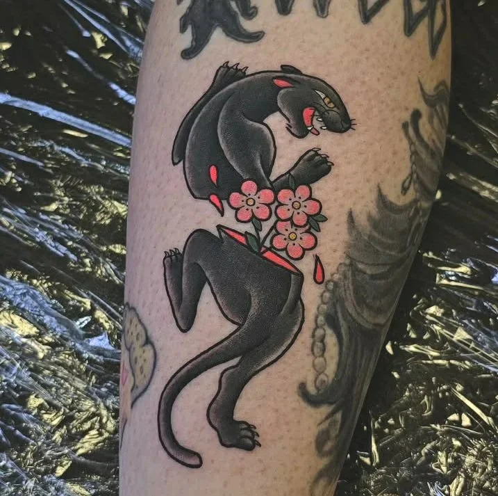 A tattoo of the American traditional crawling black panther. The panther is bisected at the waist, and three little pink flowers are growing out of it's lower half.