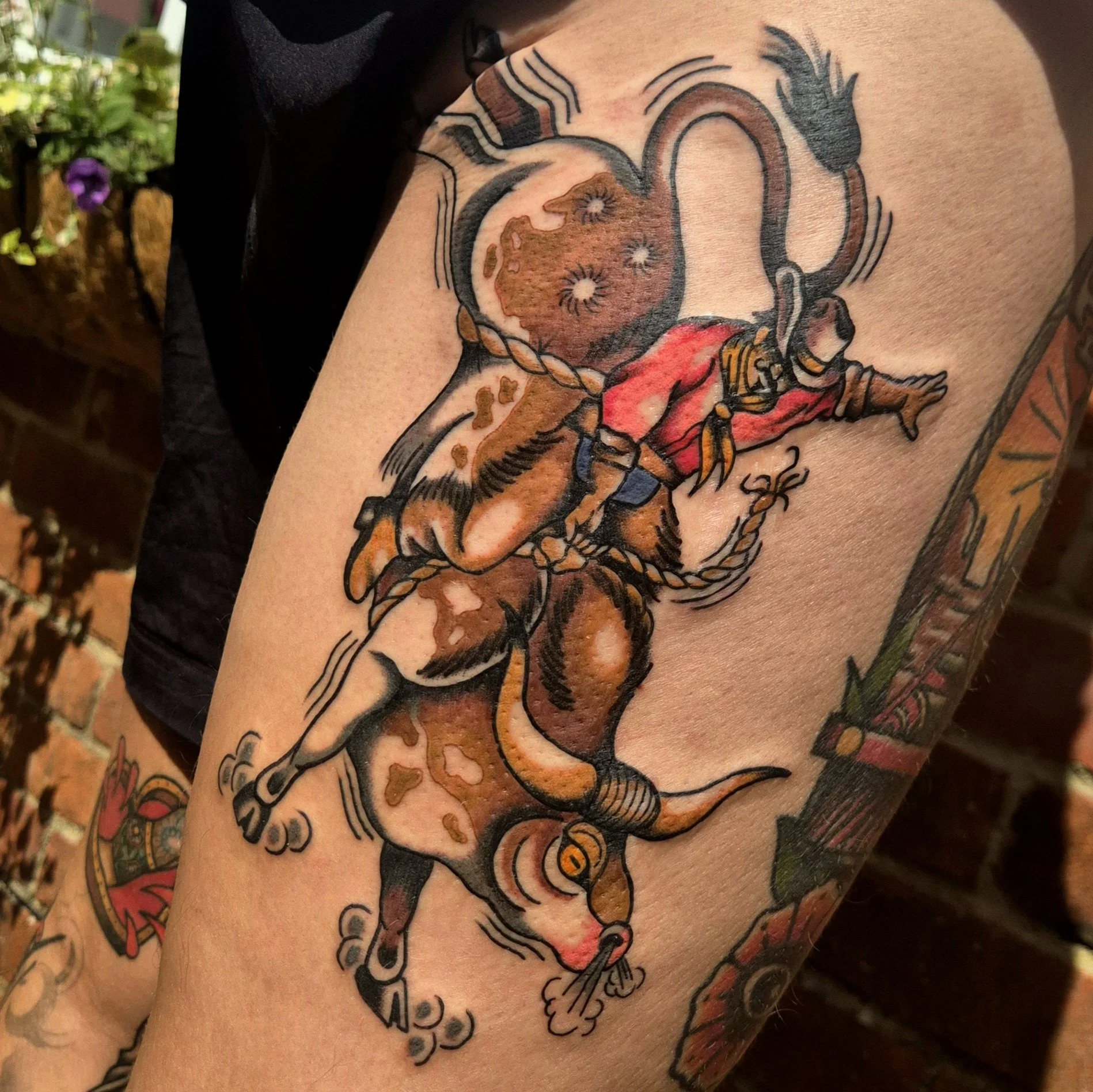 An American traditional tattoo of a cowboy riding a bull. The cowboy is wearing a red shirt, blue jeans, and brown chaps. The bull has long golden horns. The bull is bucking, but the cowboy is still riding.