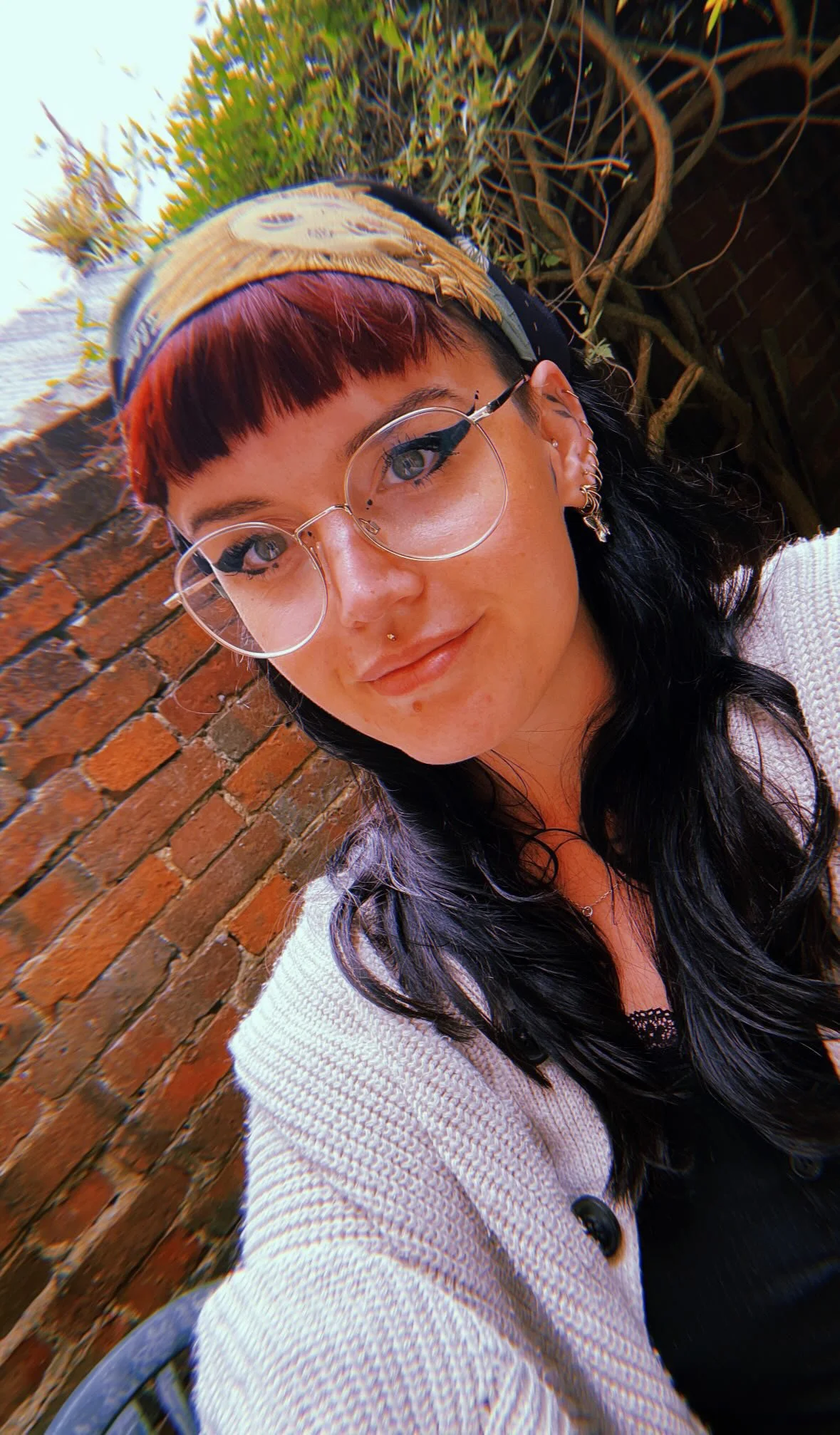 A selfie of George. She has a blunt red bang, wide square glasses with thin silver frames, and thick winged eyeliner. Her hair is long and black and she has many ear piercings. She is in front of a brick wall, and wears a white knitted shawl.