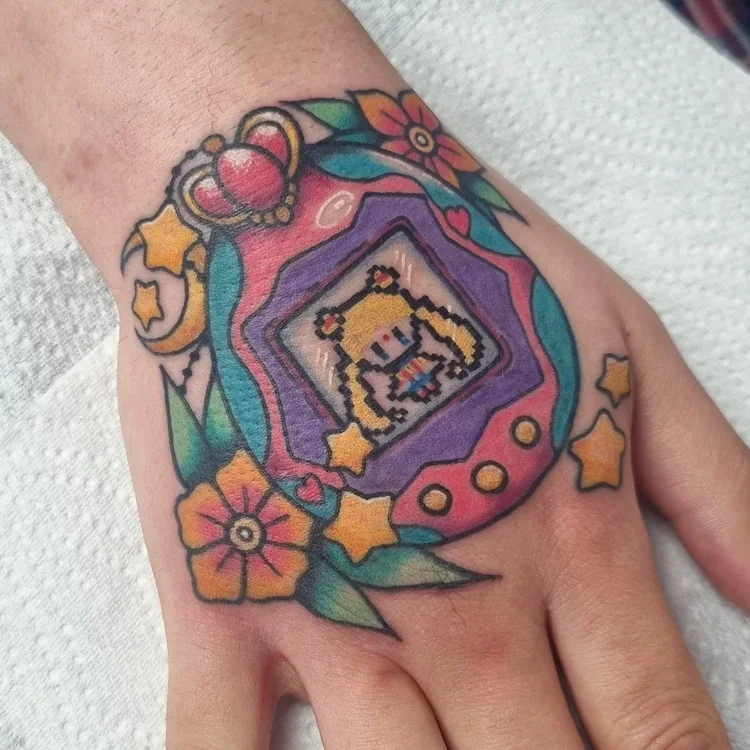 A colorful hand tattoo of a Tamagotchi toy, with a pixel art sprite of Sailor Moon on the screen. Two small orange flowers are either side of the toy, and the are several small stars and moons around it.