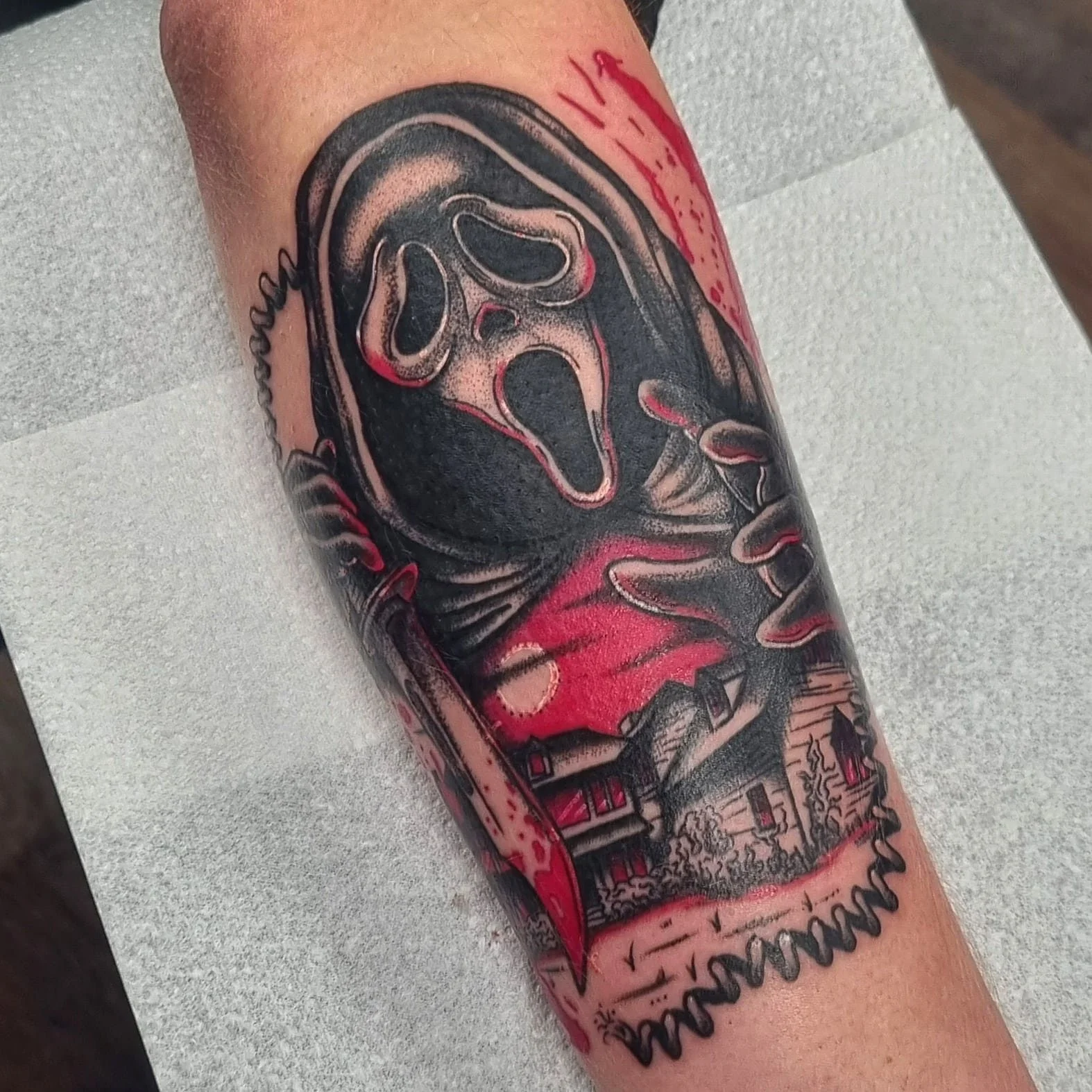 A stylized color-splash tattoo of Ghostface from the movie Scream holding a phone with one hand, reaching out over a suburban street with the other. There is a red sunset behind the house.