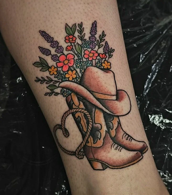 A tattoo of brown cowboy boots that are filled with colorful wild flowers. There is a brown cowboy hat resting on the boots, and a heart shaped lasso next to them.