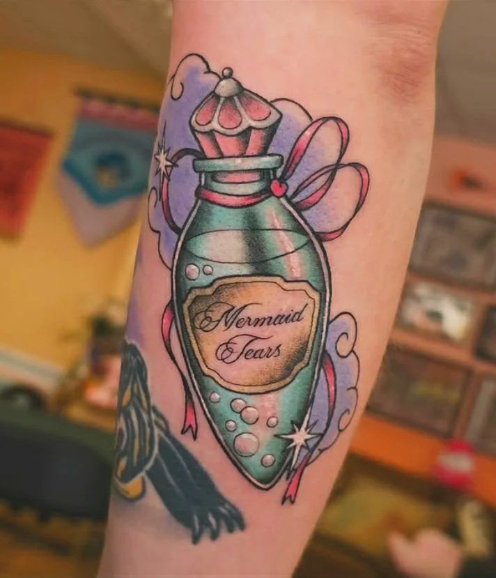 A tattoo of a ornate blue vial of liquid labeled Mermaid Tears. There is a pink ribbon around the rim, and fluffy purple clouds behind it.