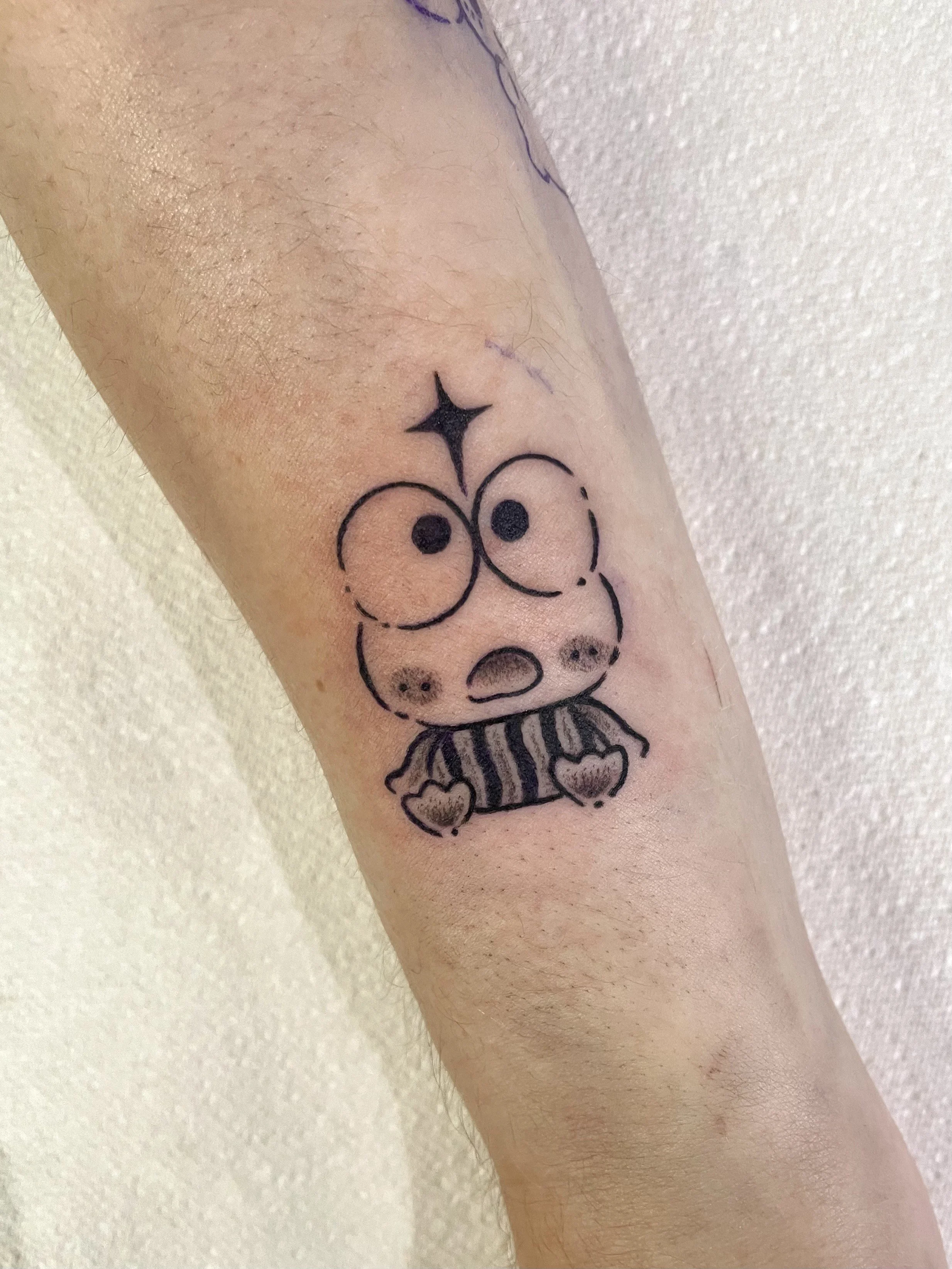 A black and white tattoo of the Sanrio character Kerrpoi. He has a surprised expression, and a little black four pointed star above his head.
