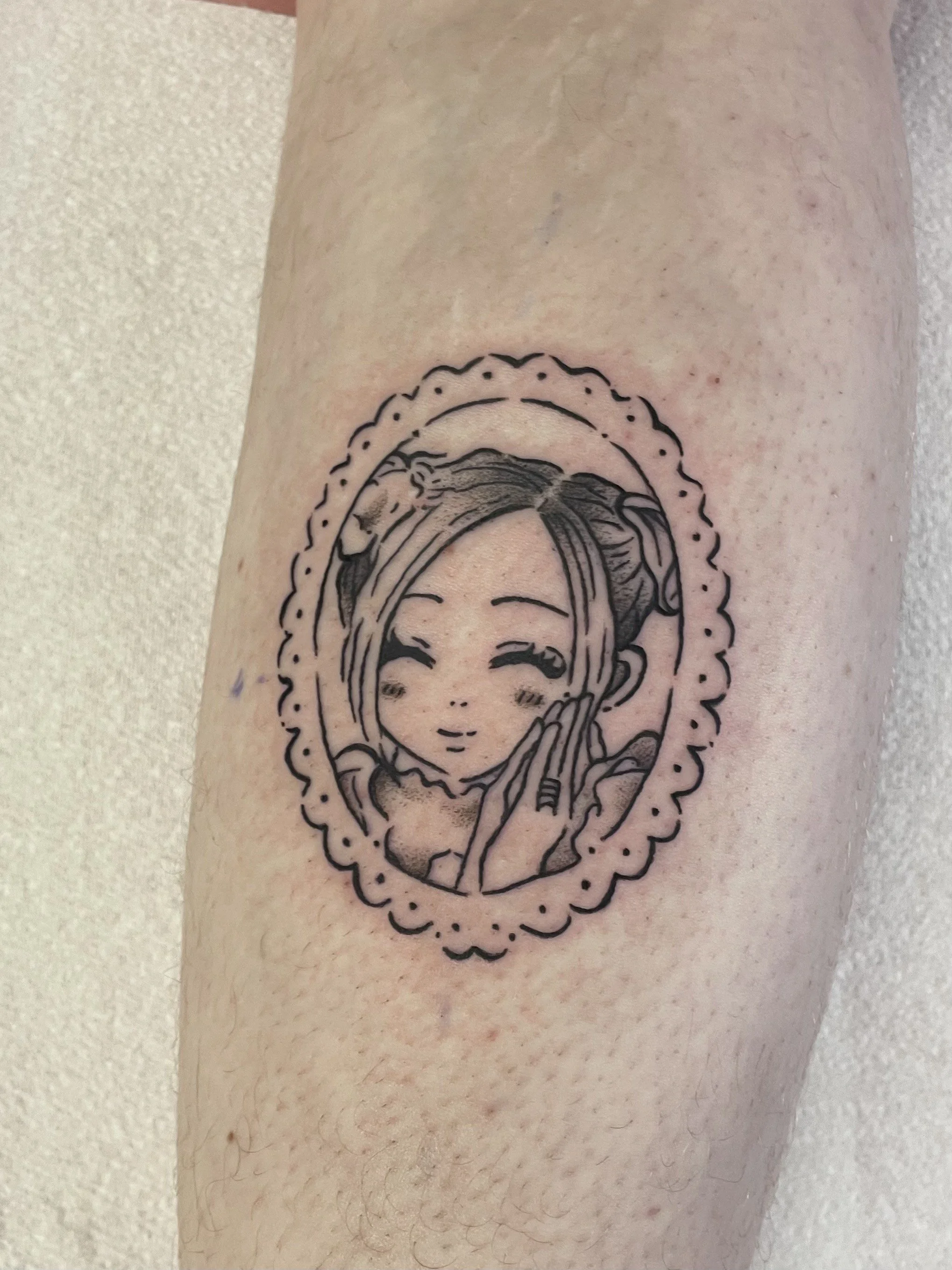 A black and white tattoo of Hachi from the anime Nana. She is smiling, and has both of her hands against her left cheek. She is in a lacey oval border.