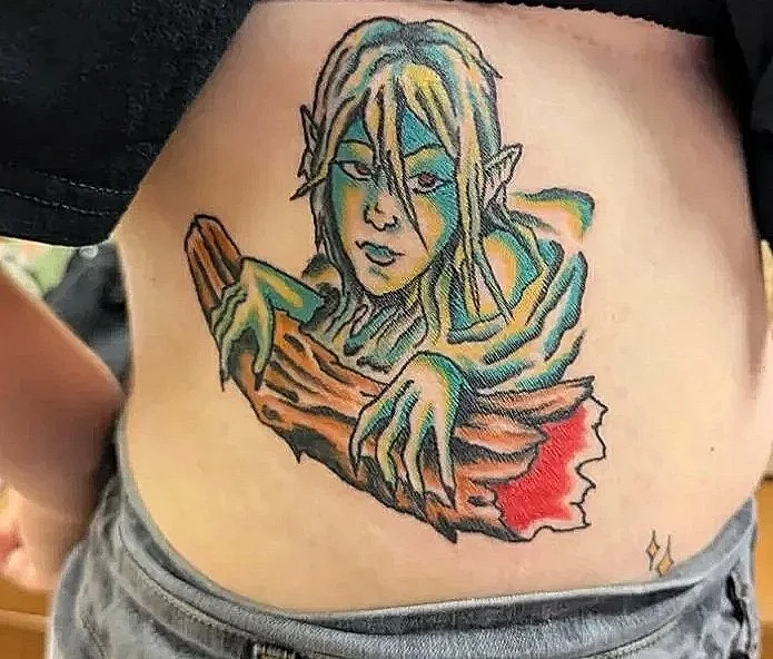 A comic style tattoo of a swamp monster woman with yellowy green skin. She is coming up out of water, and has both of her clawed hands on a brown log.