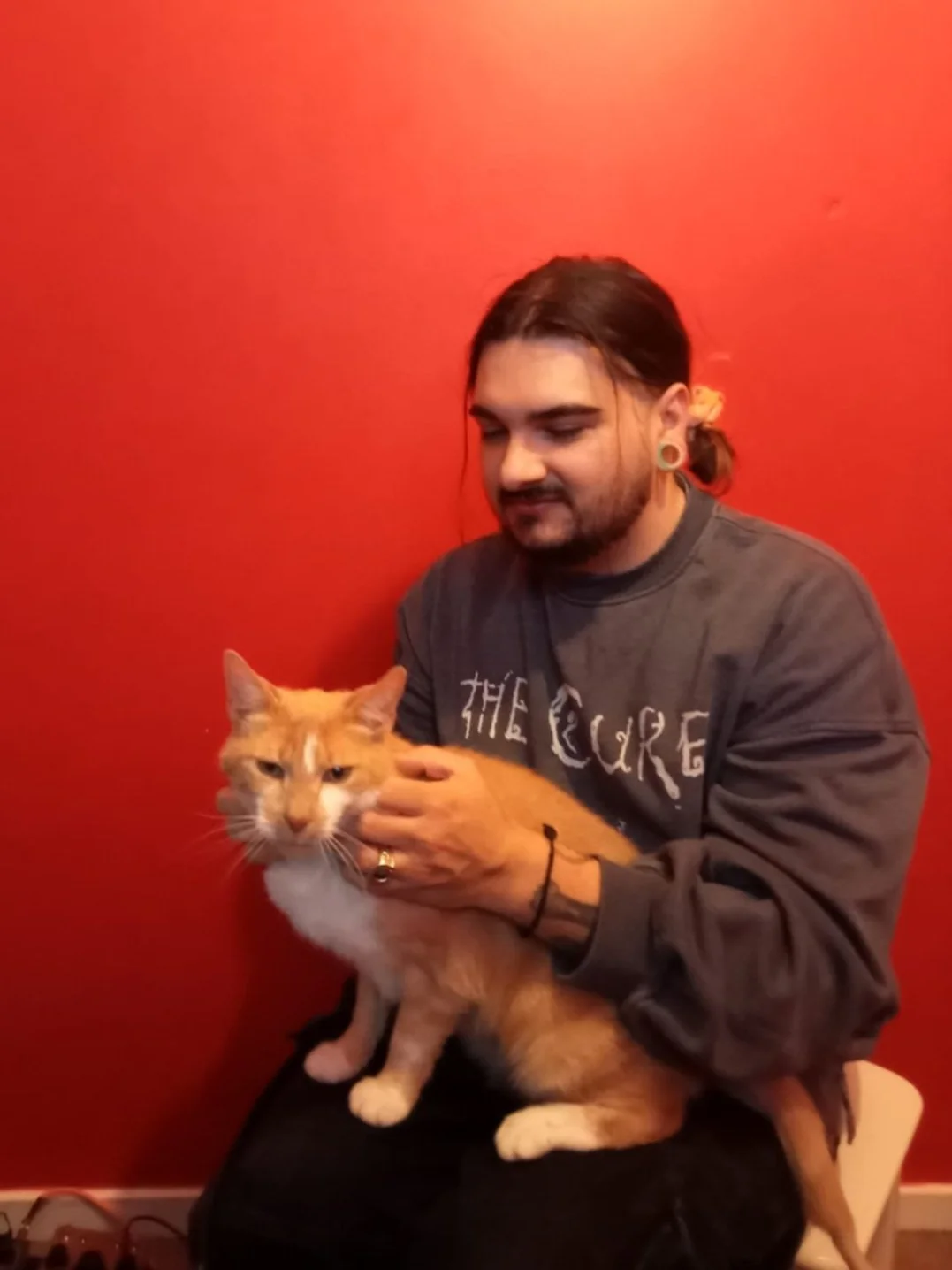 A photograph of Sam. He has long dark hair tied in a low bun. He wears a grey sweat shirt that says 'The Cure' on it. He has an orange and white cat on his lap. There is a solid red background.
