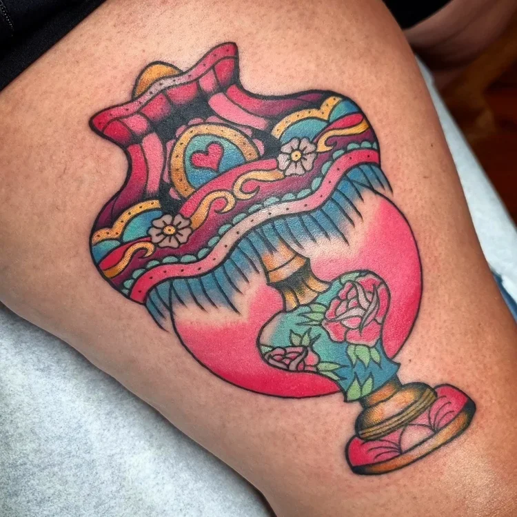 An American Traditional tattoo of an ornate lamp with matching lampshade. It is pink, blue, and gold. The base has roses on it, and the shade has a heart and two smaller flowers, as well as blue fringing.