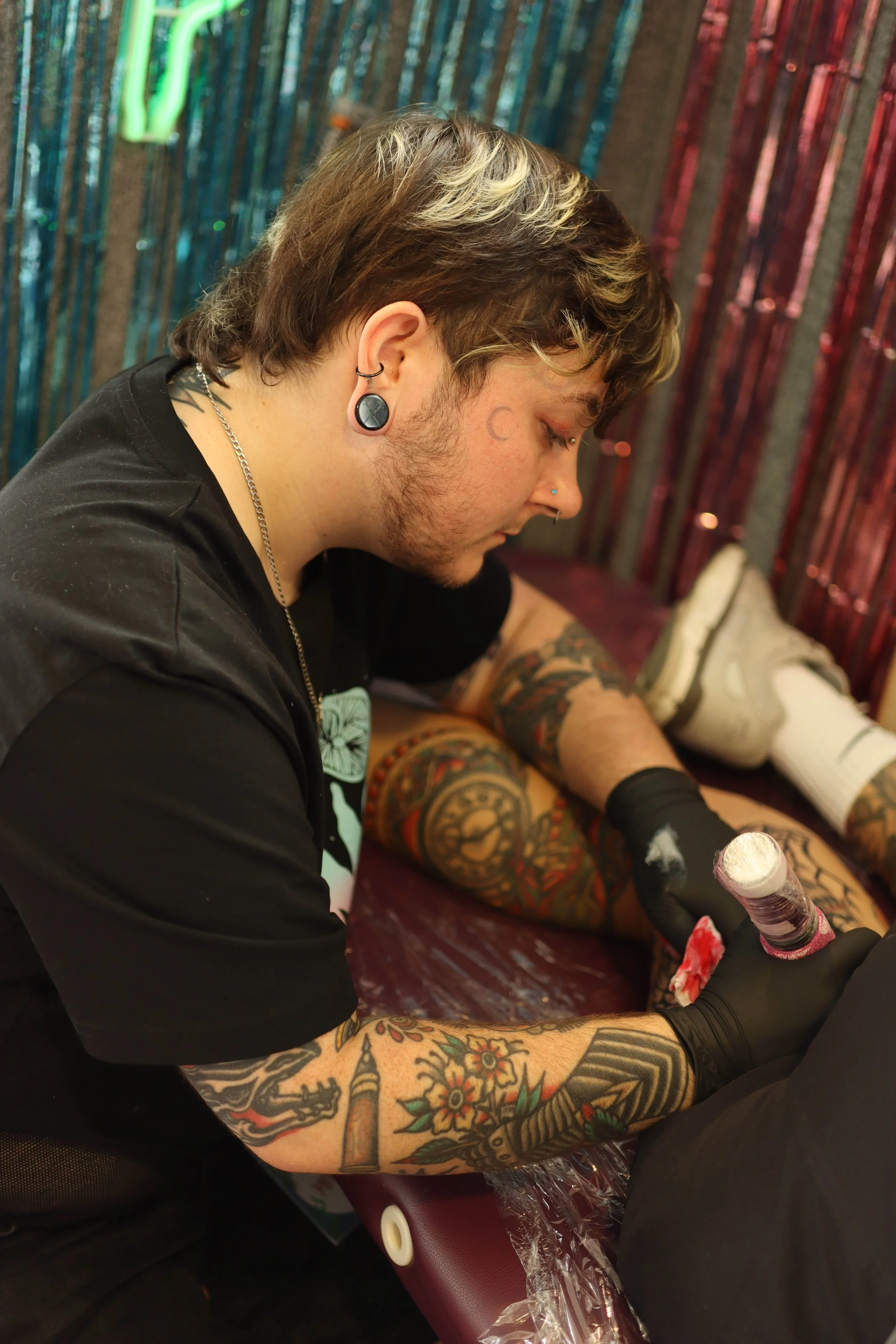 A photo of Ollie Idlehanded tattooing. He has shaggy medium brown hair with bleached chunks, a stretched lobe, and a crescent moon tattoo by his eye. He wears pink eye shadow and black gloves. The wall has pink and blue metallic fringing.
