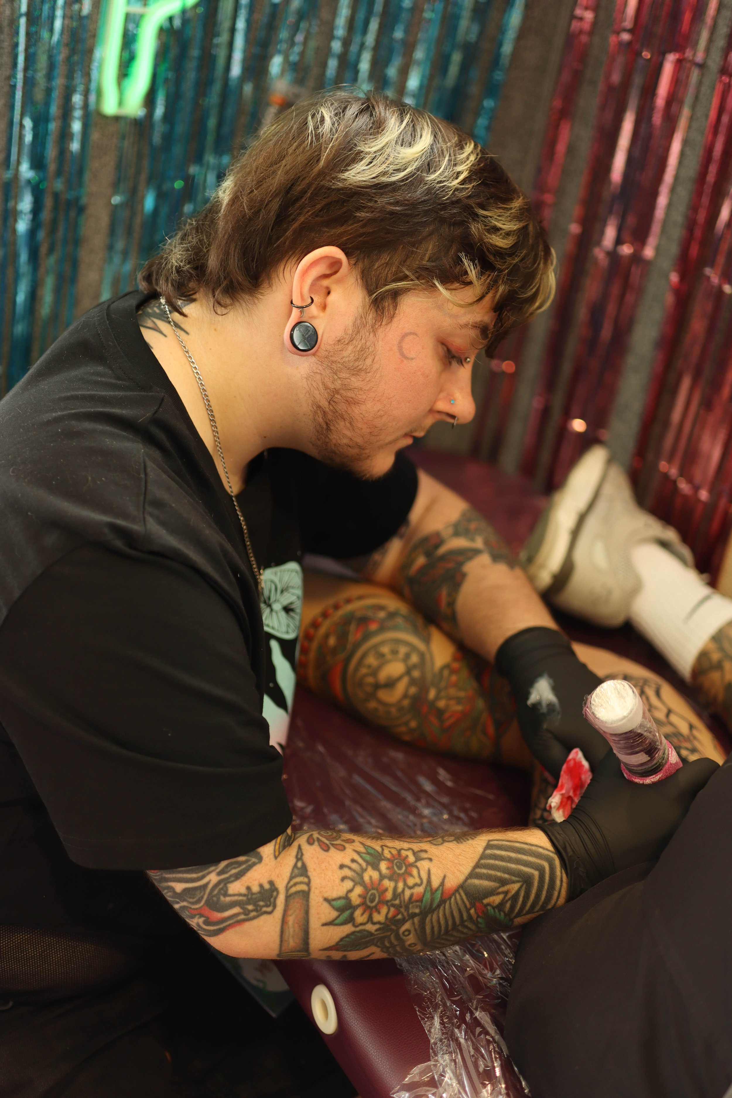 A photo of Ollie Idlehanded tattooing. He has shaggy medium brown hair with bleached chunks, a stretched lobe, and a crescent moon tattoo by his eye. He wears pink eye shadow and black gloves. The wall has pink and blue metallic fringing.