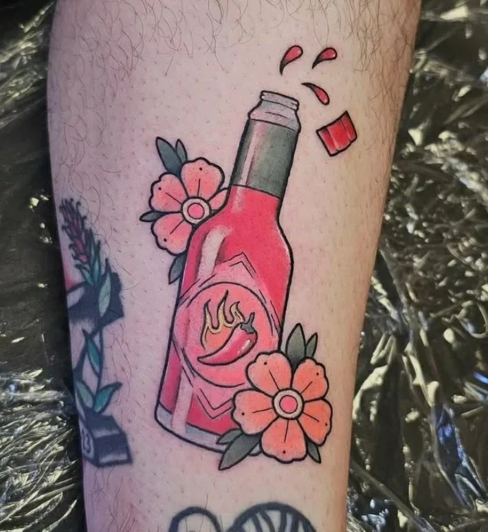 A tattoo of a hot sauce bottle. The bottle resembles a Tobasco bottle - it is red, with a green stripe on the neck. It features a flaming chili pepper on the front. Two orange flowers are next to the bottle, and the cap is popped off.