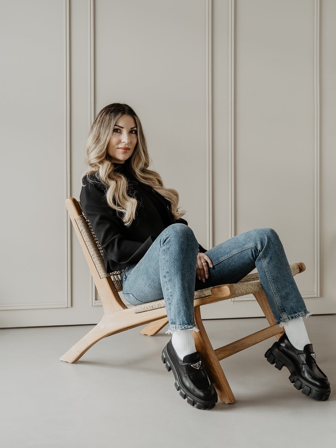 Woman sitting on a wooden chair against a beige paneled wall, wearing a black blazer, jeans, white socks, and black chunky shoes.