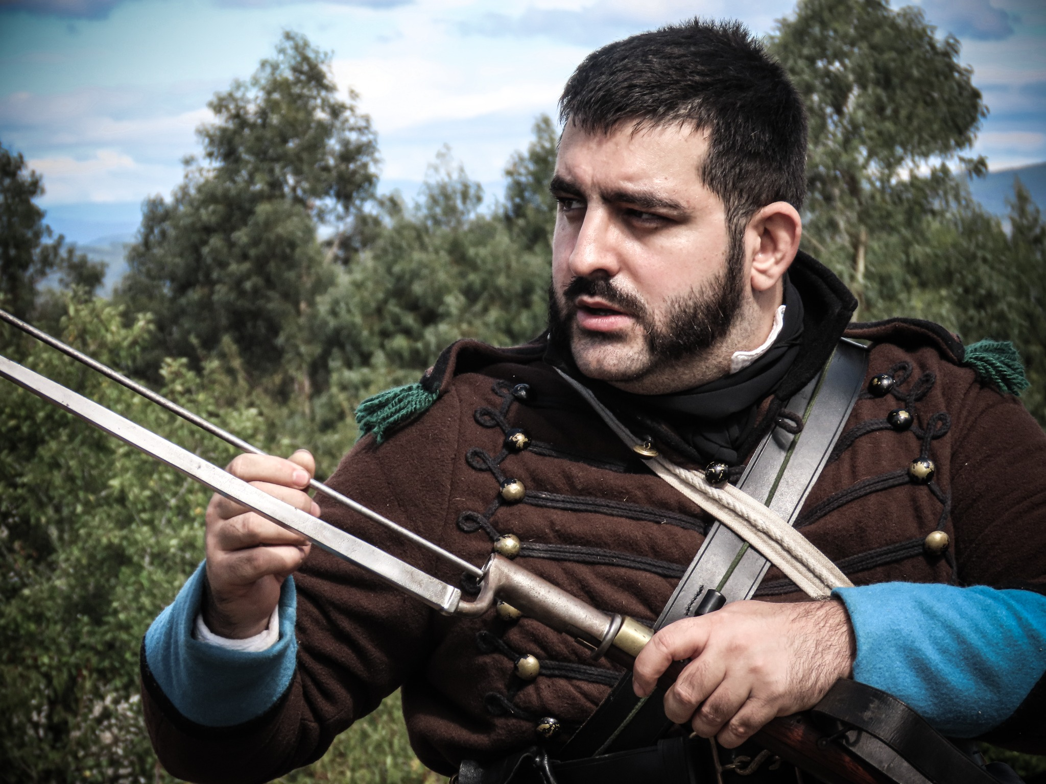 A man dressed in historical military attire, holding a musket, standing outdoors with trees and a cloudy sky in the background.