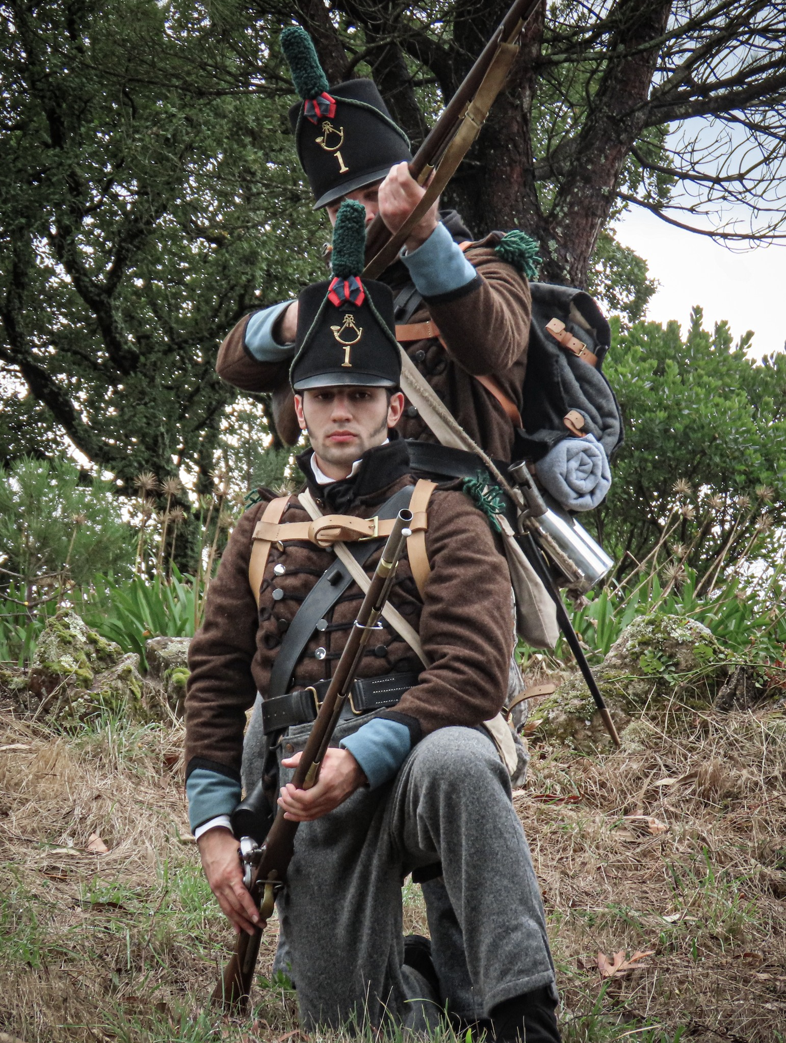 Two men dressed as 19th-century soldiers, wearing brown coats and black hats with a gold emblem and a number 1. They are outdoors in a wooded area with grass and plants, holding rifles, with one kneeling and the other standing behind him.