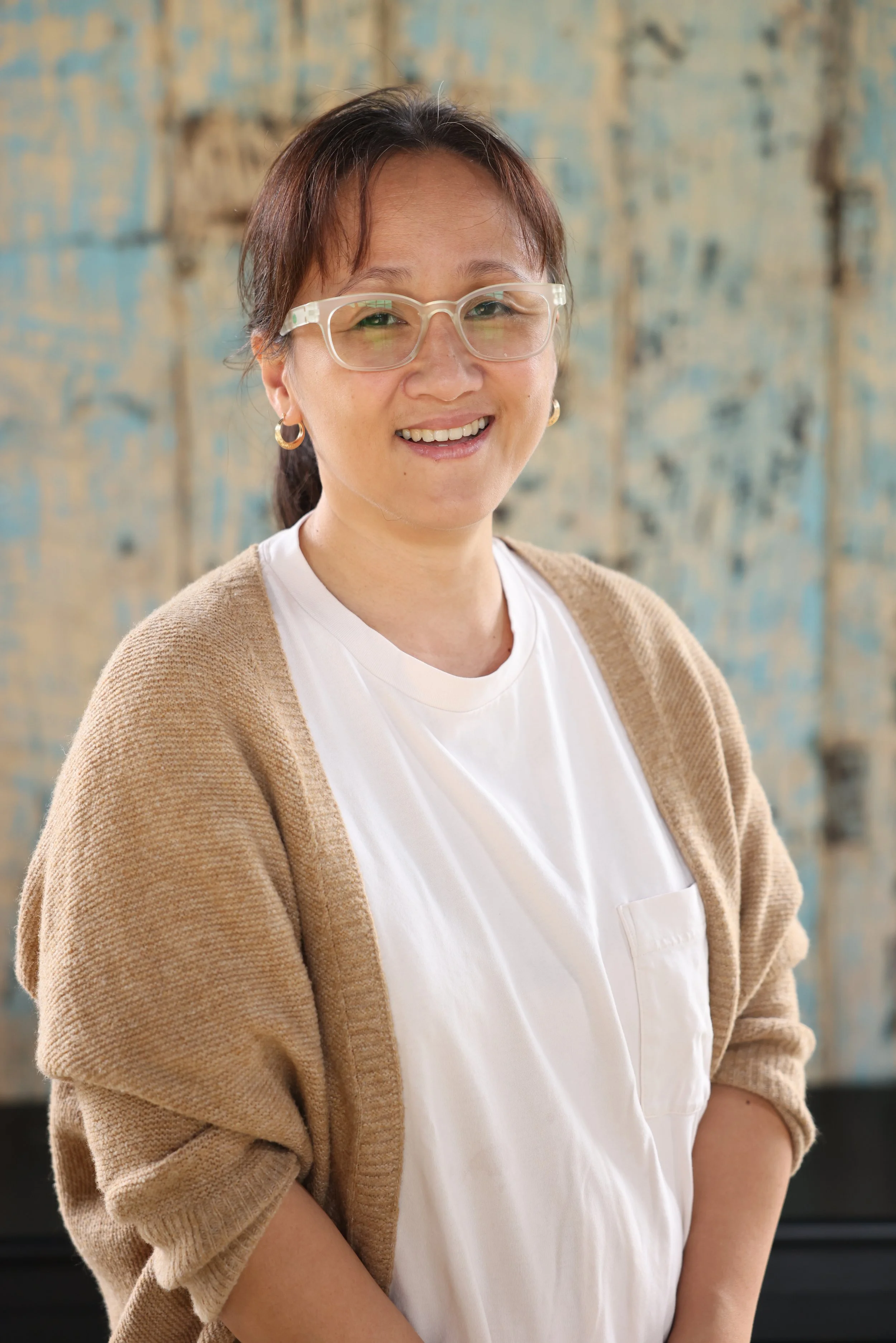 Smiling person wearing glasses, a white shirt, and a beige cardigan, standing in front of a textured blue and beige background.