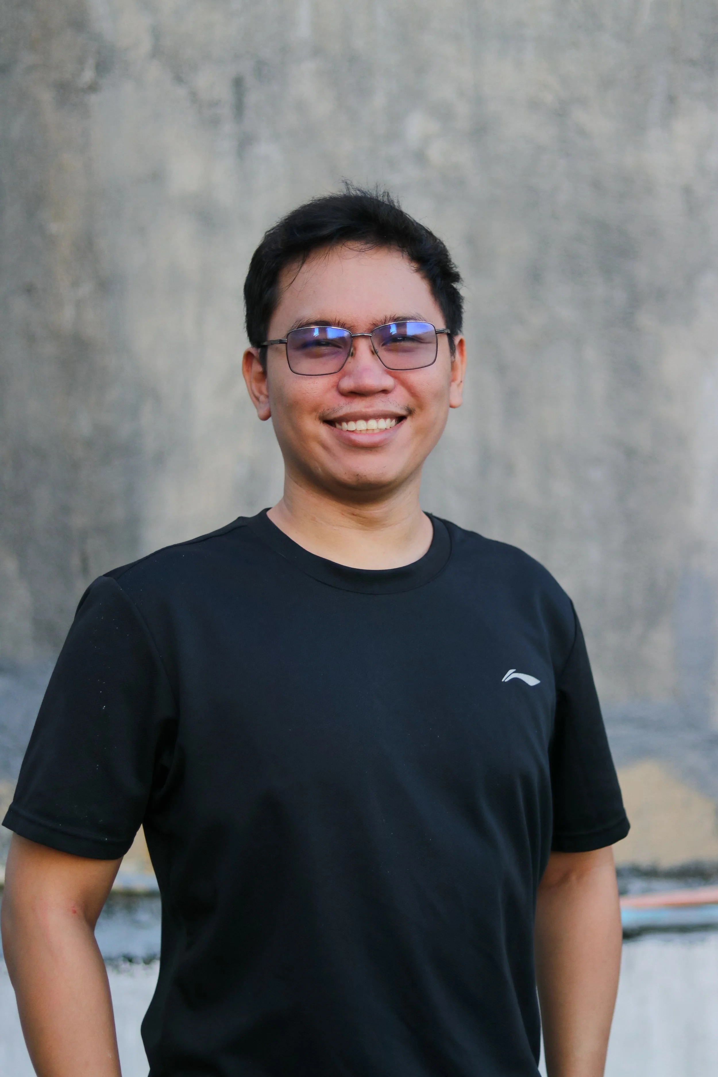 Smiling person wearing glasses and a black t-shirt standing outdoors, with a textured wall in the background.