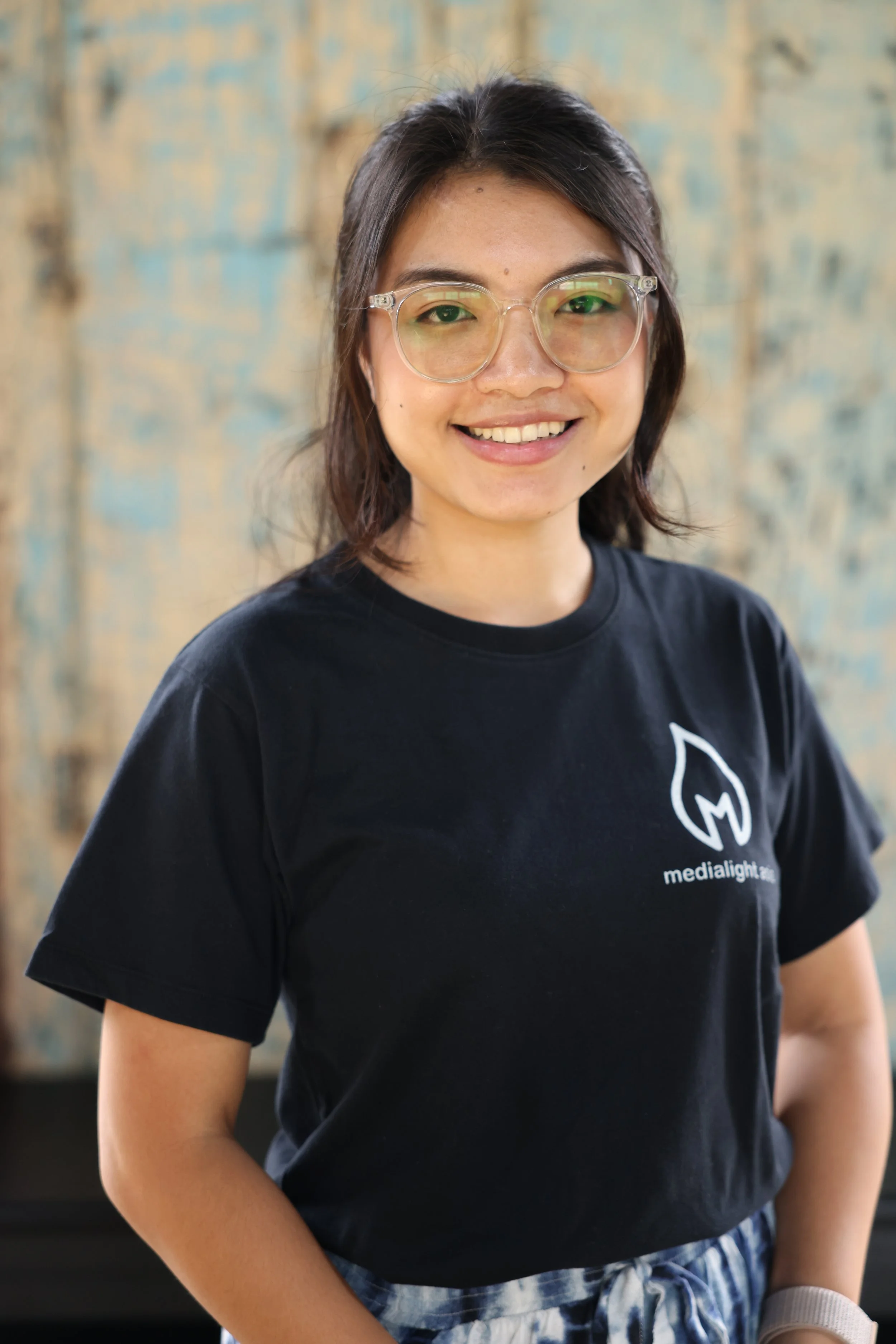 Person wearing glasses and a black Mediagrill.com t-shirt, smiling in front of a textured background.