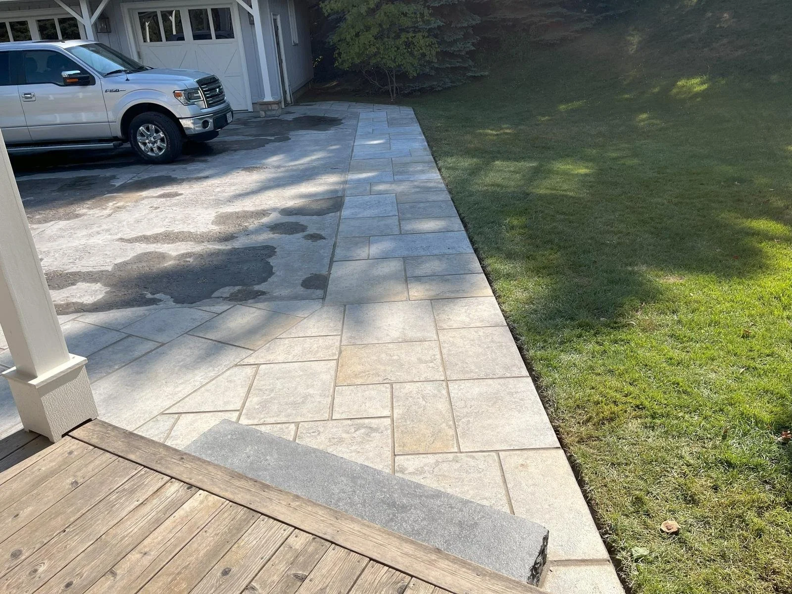 A driveway with a silver pickup truck parked on it, adjacent to a stone patio and a grassy lawn, with a white garage door and trees in the background.